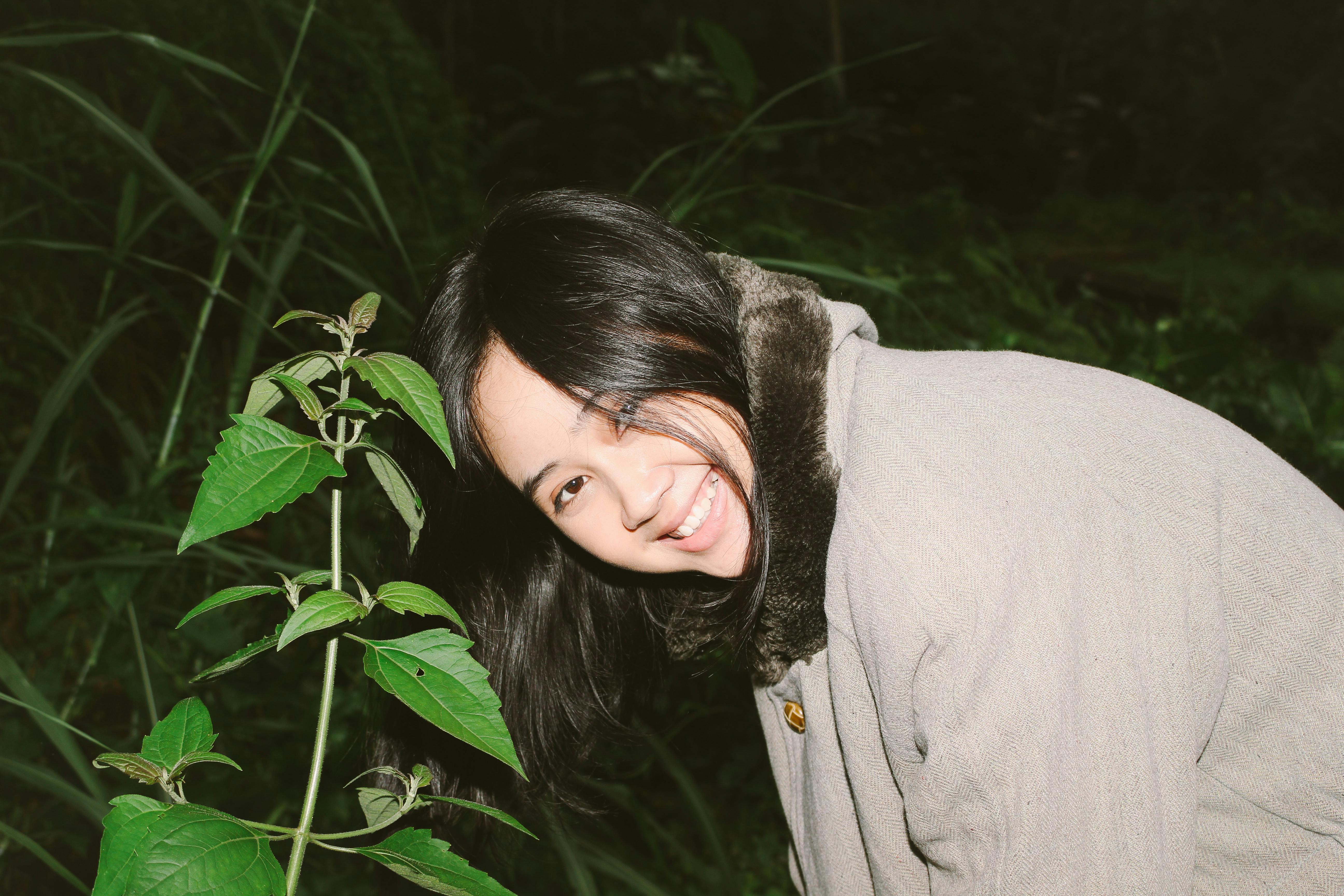 Ilmainen Iloinen hymy huulilla ja mukavassa takissa oleva nainen on ulkona yöllä rehevien vihreiden kasvien lähellä. Kuvapankkikuva