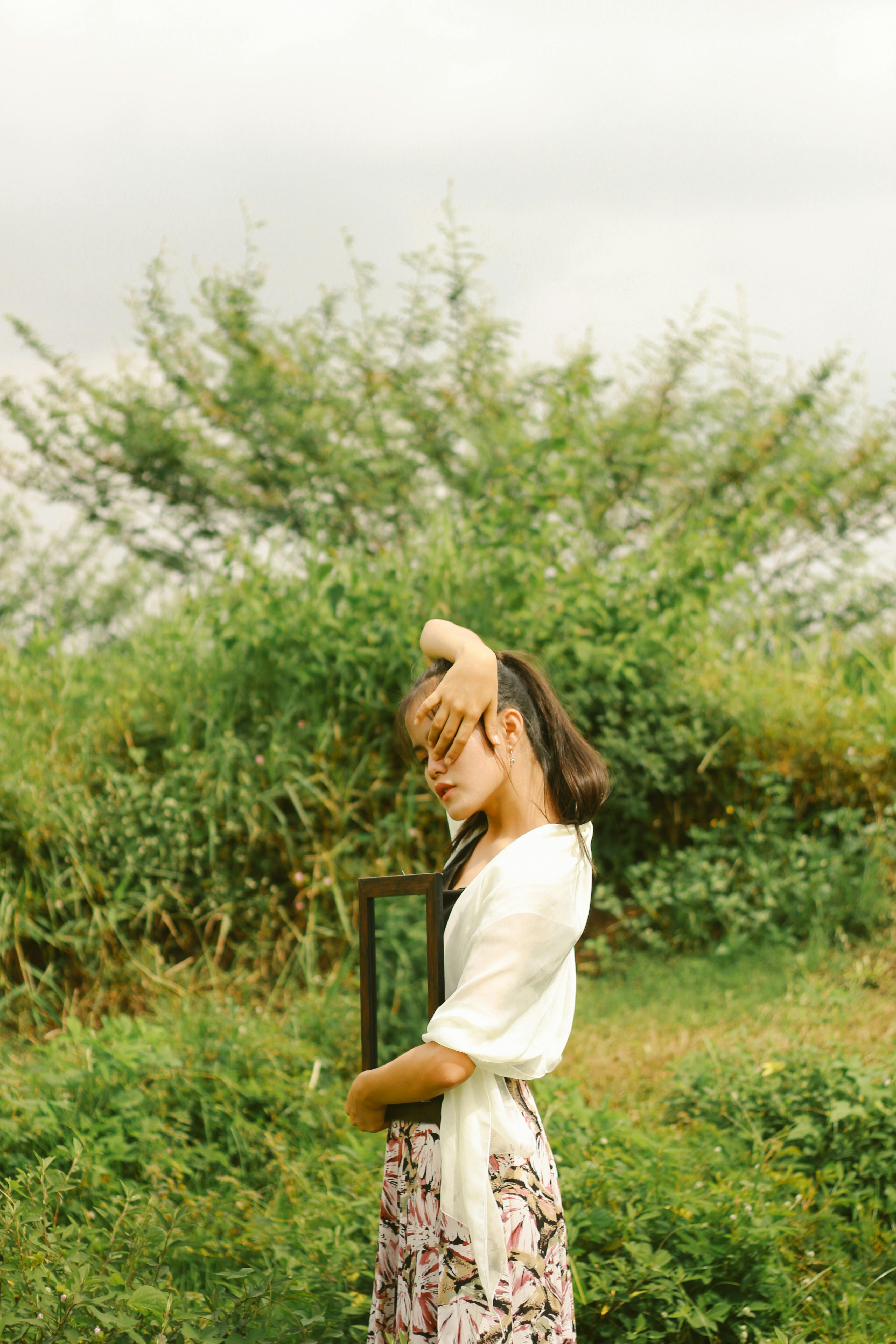 Gratis Potret artistik seorang wanita muda dengan bingkai di tengah hamparan ladang hijau yang semarak, membangkitkan kreativitas dan introspeksi. Foto Stok