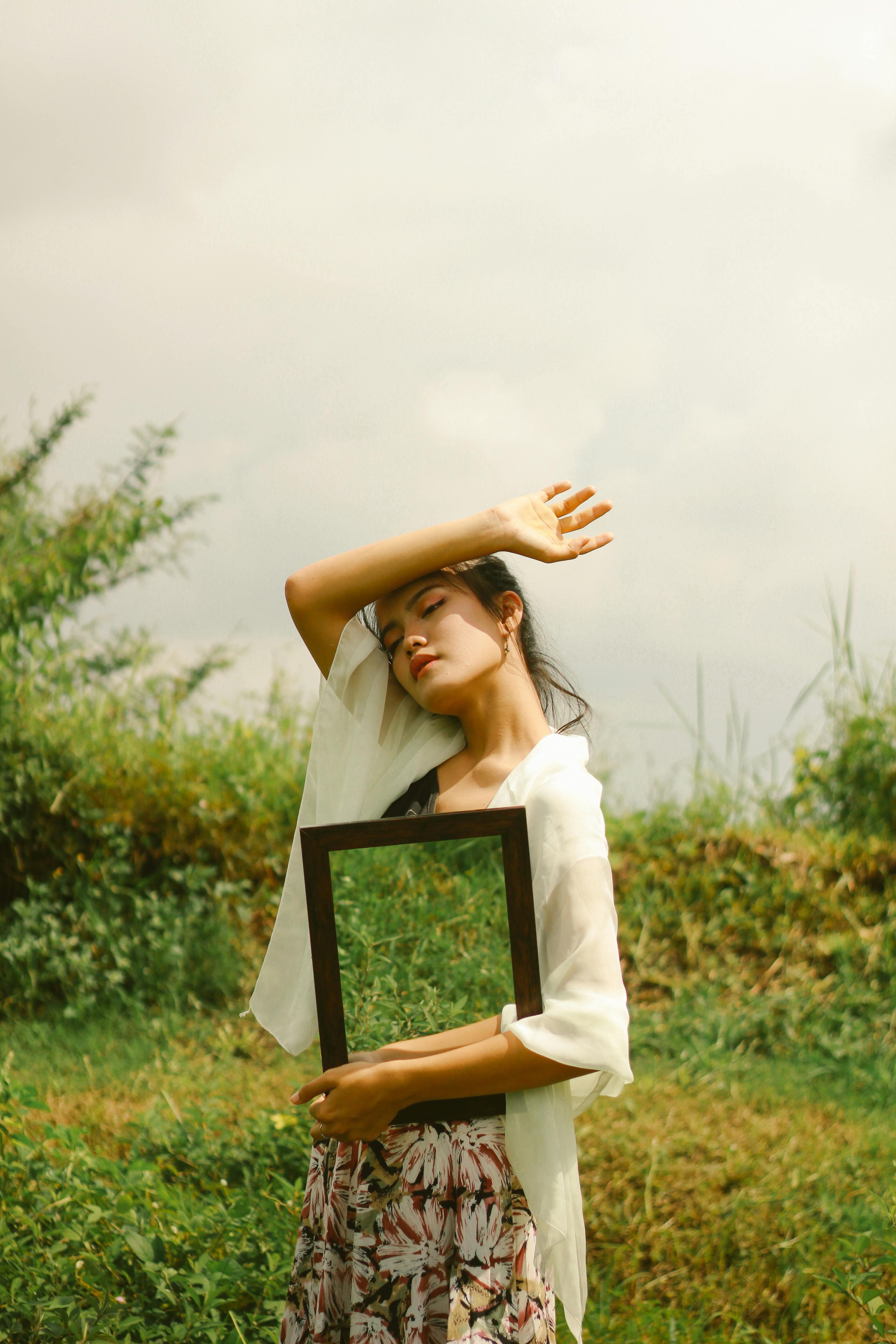 An artistic portrait of a woman holding a frame, set in a serene lush landscape.