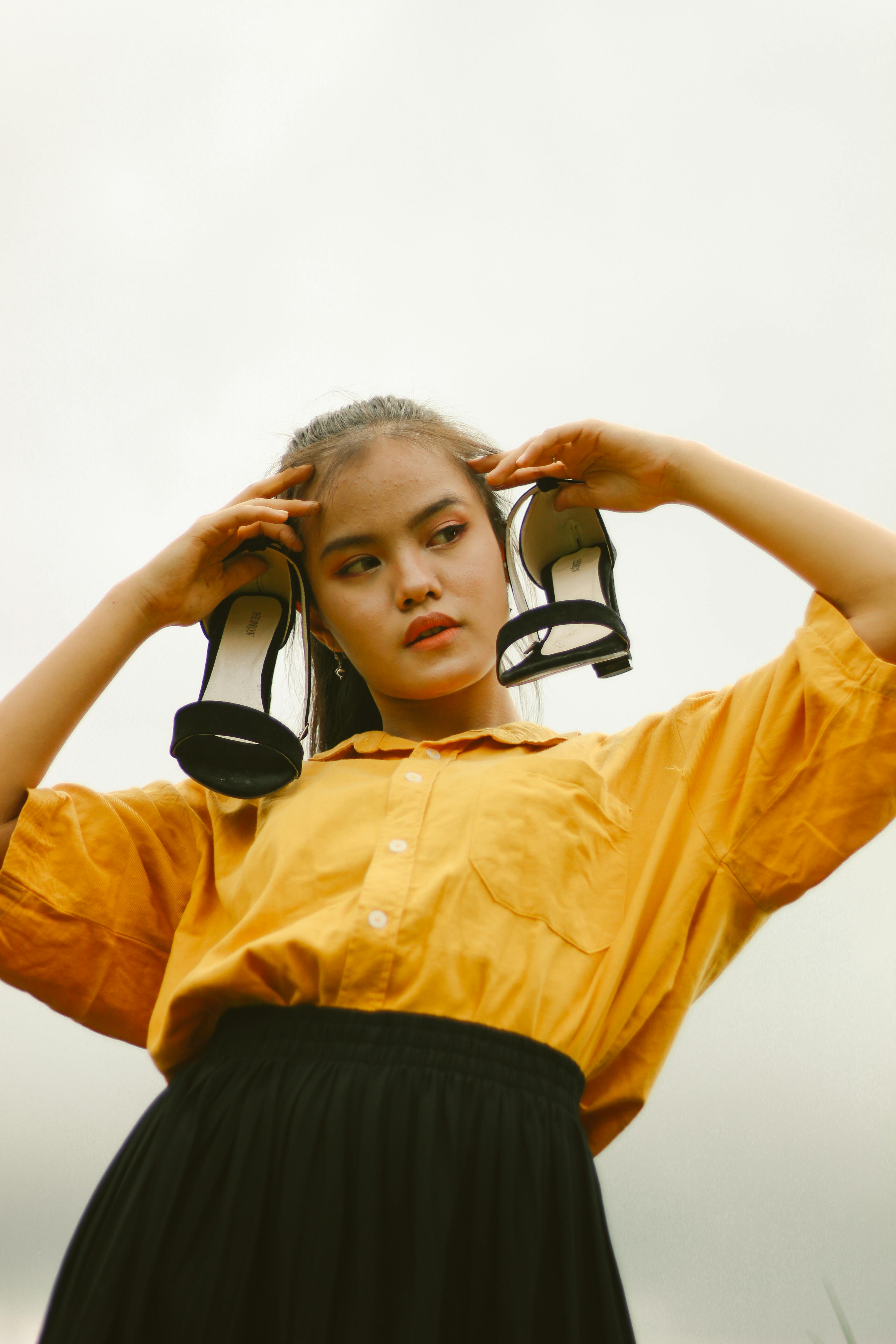 Miễn phí Một cô gái trẻ tạo dáng ngoài trời với đôi giày cao gót đặt trên vai, thể hiện sự sáng tạo trong thời trang. Ảnh lưu trữ