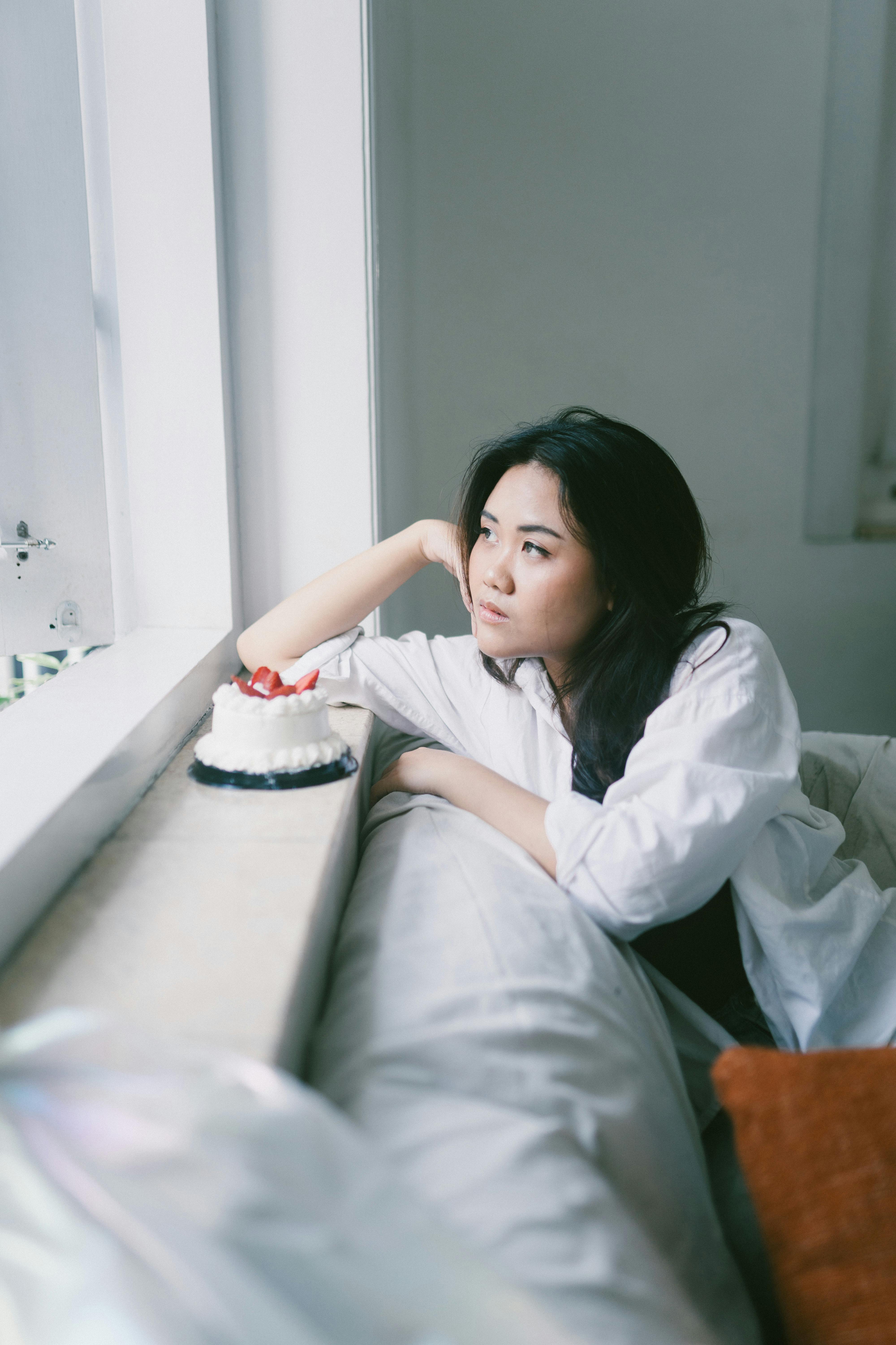 bezplatná Zamyšlená žena se zamyšleně dívá z okna vedle malého dortu. Základová fotografie