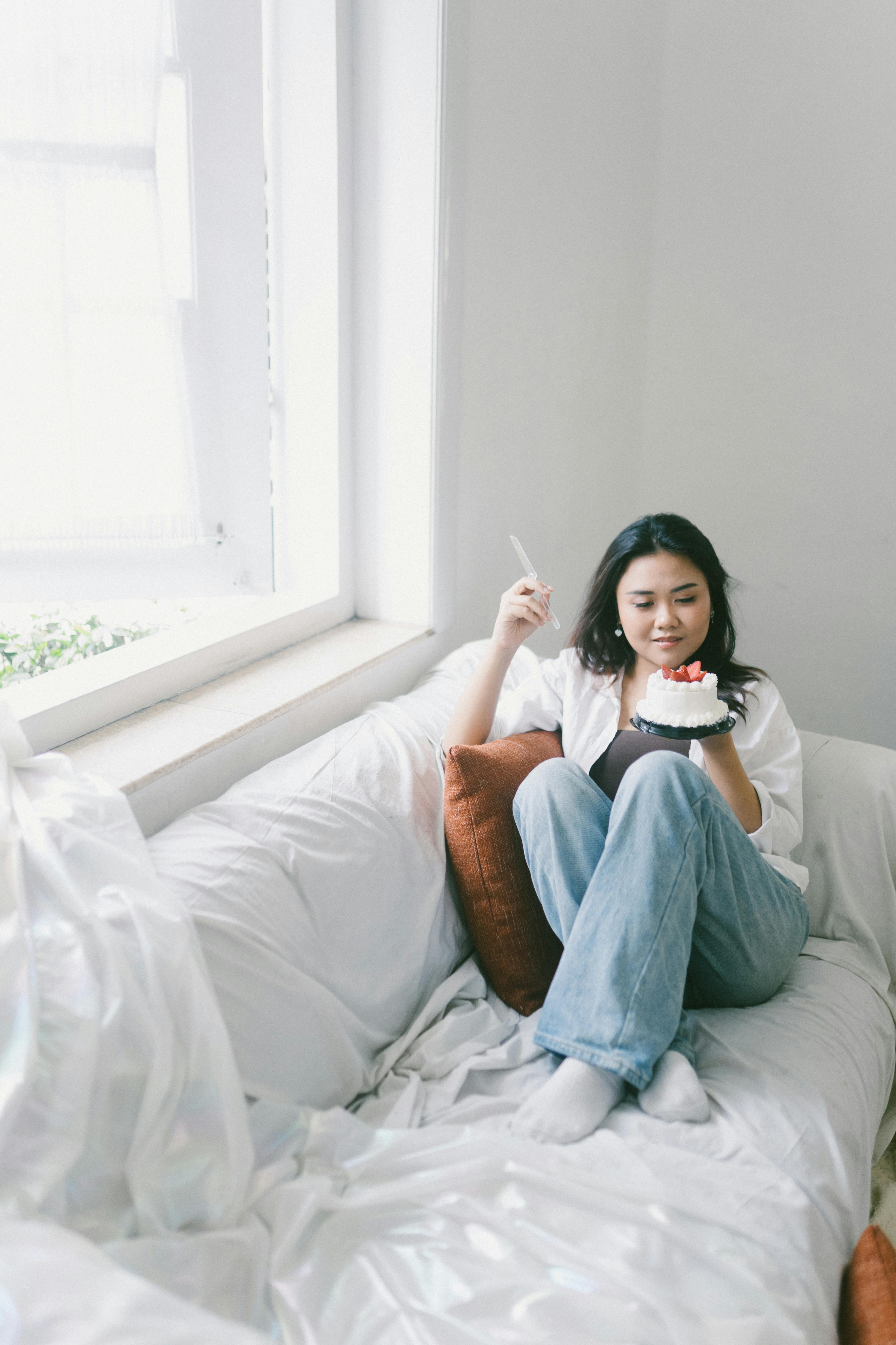 Bezpłatne Młoda kobieta siedzi na łóżku z kawałkiem ciasta w jasnym, przytulnym pokoju. Zdjęcie z galerii