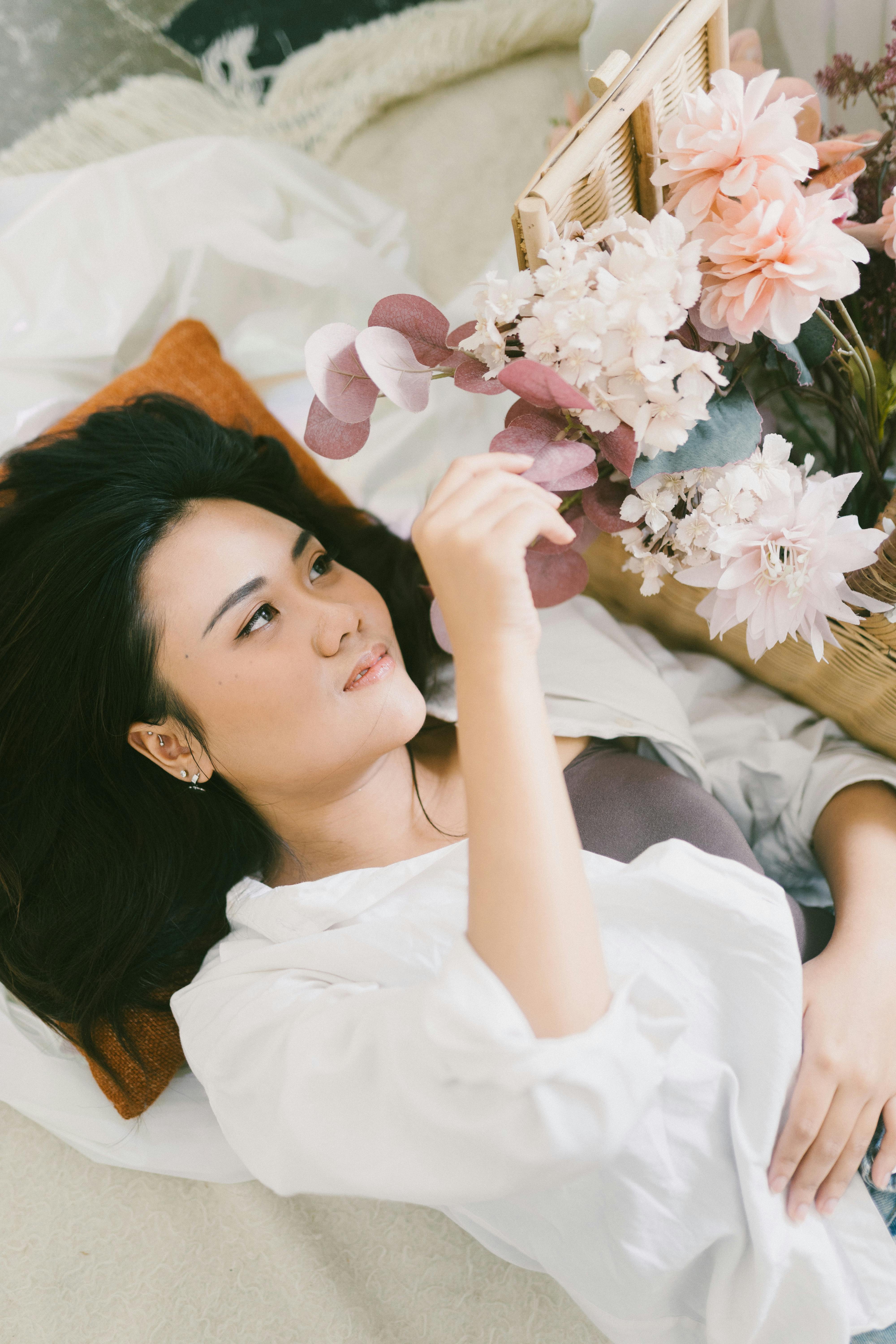 Bezpłatne Kobieta leżąca na łóżku, trzymająca w ręku i podziwiająca kolorowy bukiet kwiatów w pomieszczeniu. Zdjęcie z galerii