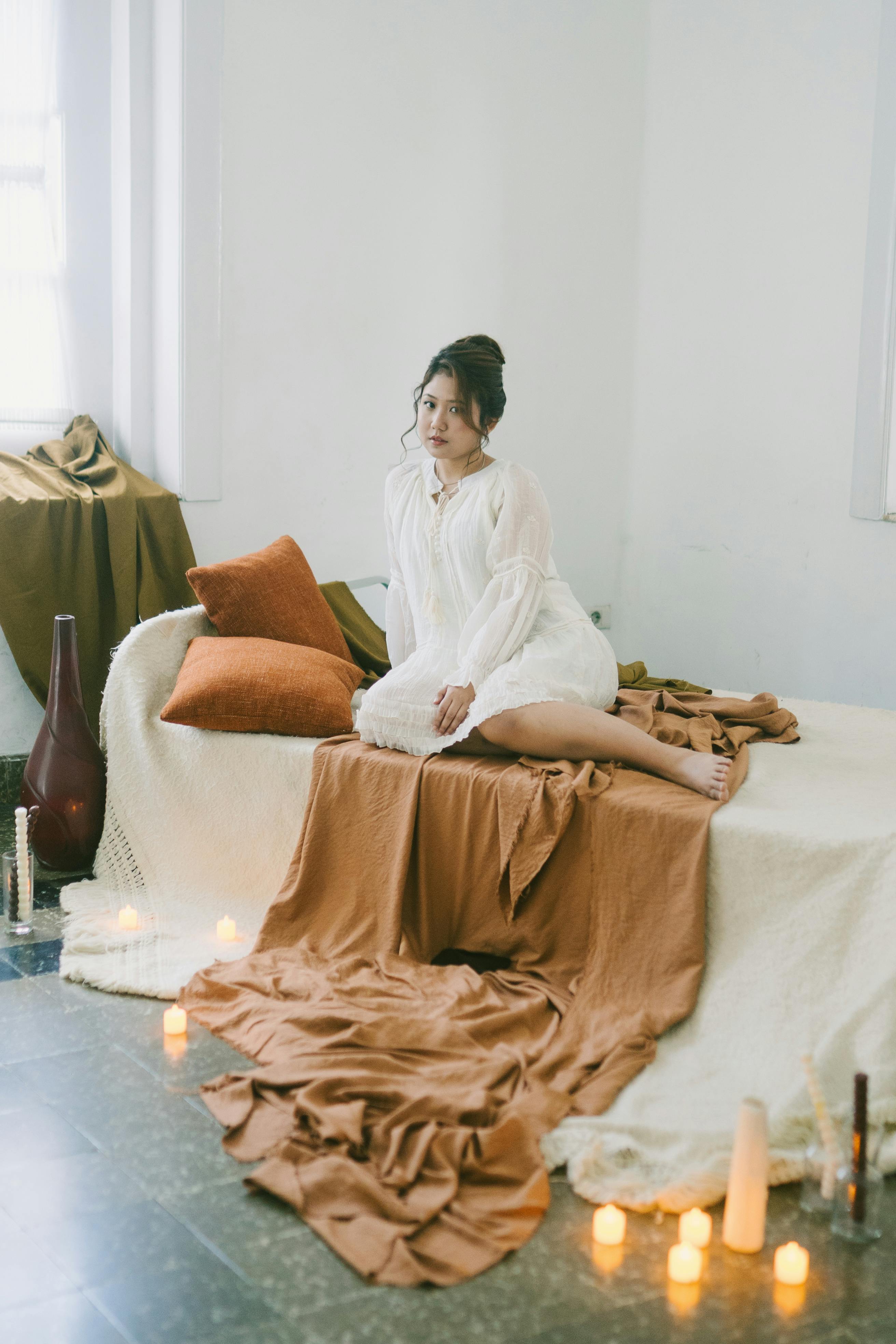 Miễn phí Người phụ nữ thanh lịch trong chiếc váy trắng ngồi trên giường với những ngọn nến, toát lên vẻ bình yên và ấm áp trong một khung cảnh ấm cúng. Ảnh lưu trữ