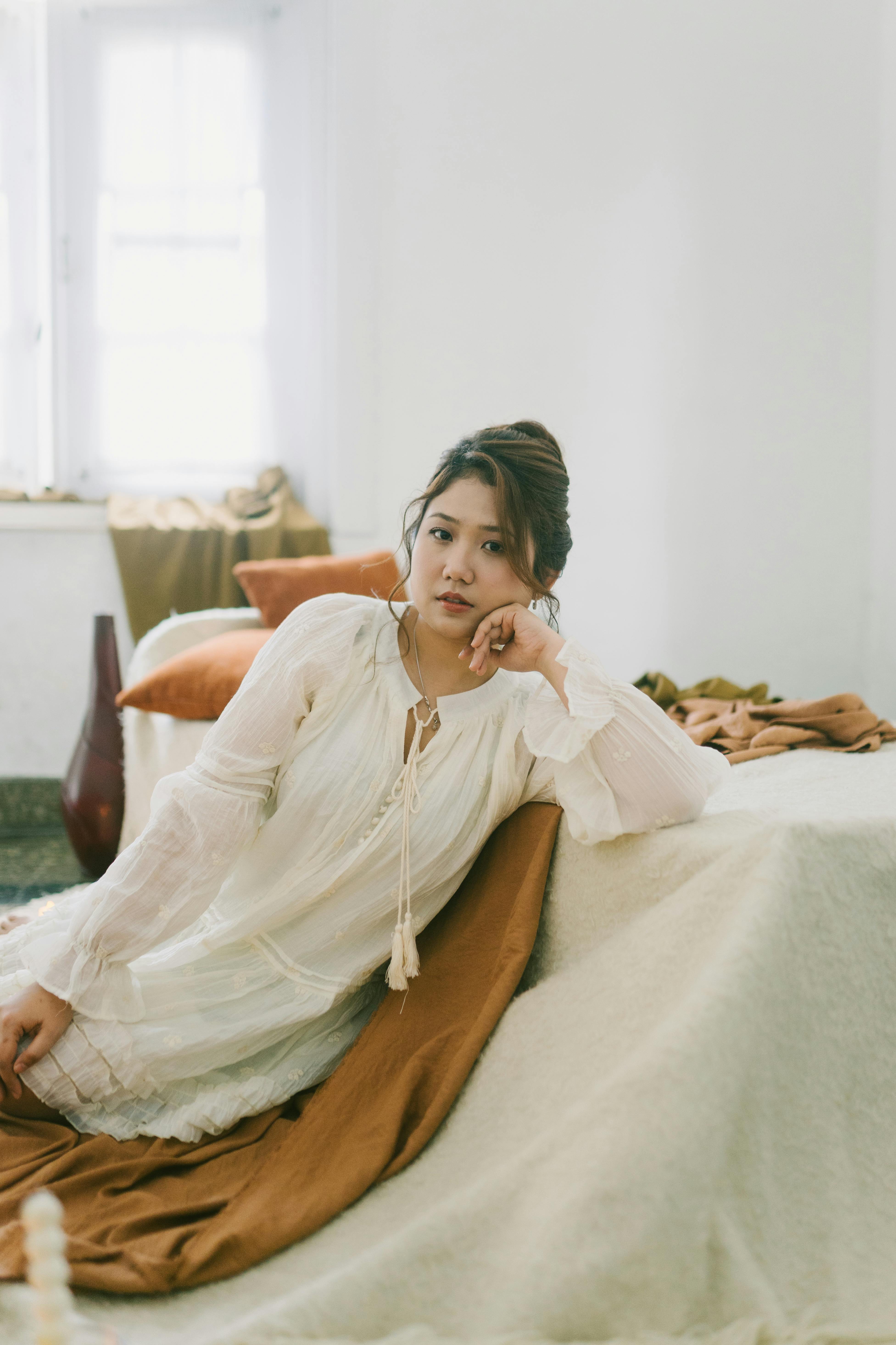 Miễn phí Một người phụ nữ mặc váy trắng đang ngồi trên giường trong một căn phòng được chiếu sáng dịu nhẹ. Ảnh lưu trữ