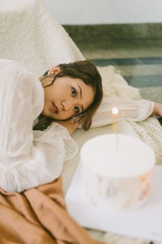 A young woman lying indoors beside a white birthday cake with a lit candle.