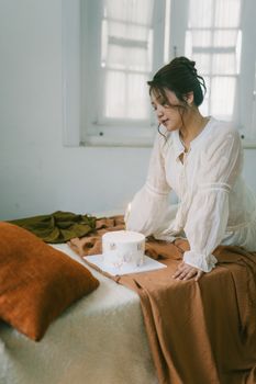 A woman in a white dress contemplates a candlelit cake in a cozy, softly lit room.