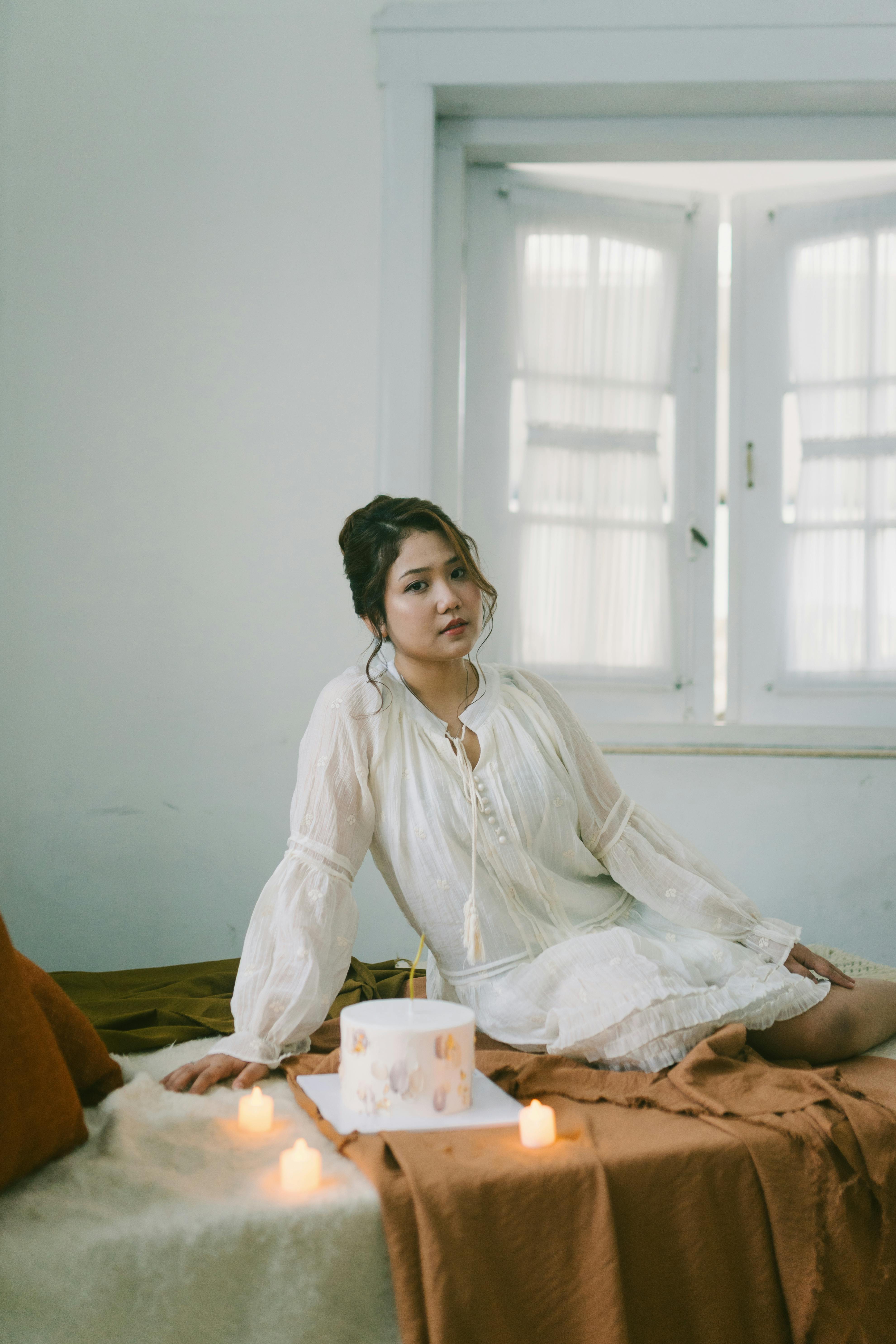 bezplatná Mladá žena v bílých šatech sedí v útulném pokoji osvětleném svíčkami a měkkým světlem, z něhož vyzařuje klid. Základová fotografie