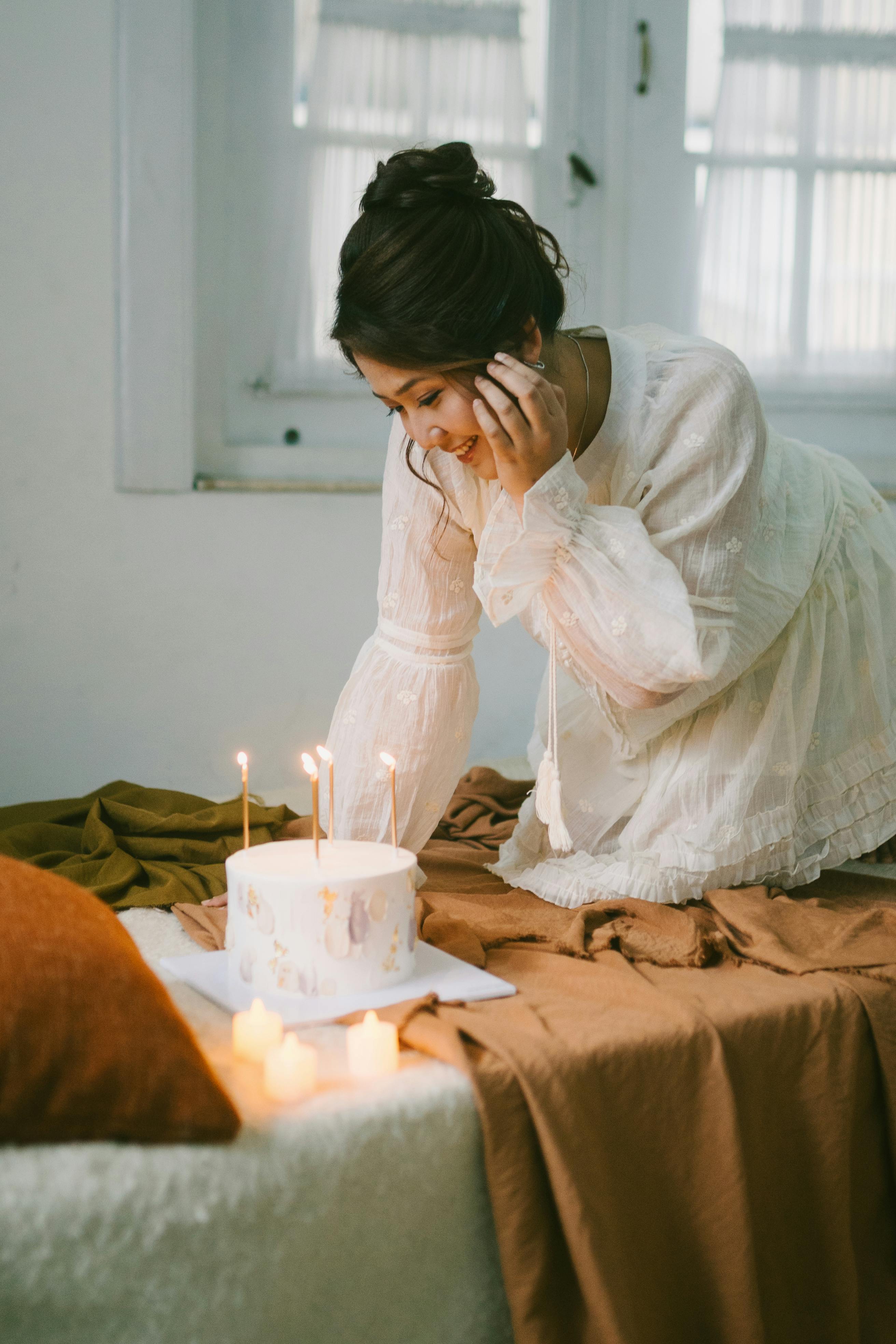 Ücretsiz Beyaz elbiseli bir kadın, üzerinde mumlar yakılmış doğum günü pastasının tadını çıkarıyor ve bu da sıcak bir atmosfer yaratıyor. Stok Fotoğraflar