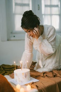A young woman in a white dress leans over a birthday cake with candles in an intimate setting.