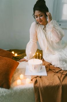 Woman in white dress celebrating birthday with candle-lit cake in cozy indoor setting.