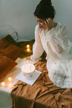 Young woman in a white dress celebrating a cozy birthday indoors with candles and a cake.