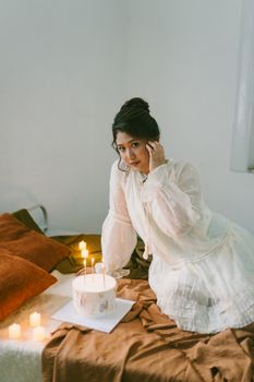 Woman in white dress celebrates birthday in cozy room with cake and candles creating a warm atmosphere.