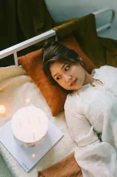 Relaxed young woman lying on bed beside a candlelit cake, creating a warm, cozy atmosphere.