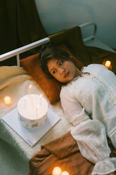 A woman in a white dress is lying on a bed surrounded by candles and a cake, creating a cozy atmosphere.