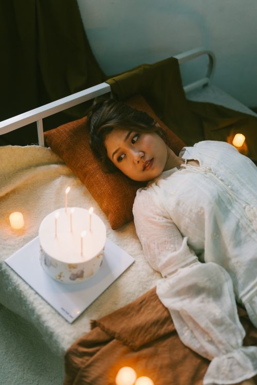 Free A woman in a white dress is lying on a bed surrounded by candles and a cake, creating a cozy atmosphere. Stock Photo