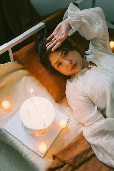 Woman in white blouse resting beside a cake and candles, creating a serene and cozy atmosphere.
