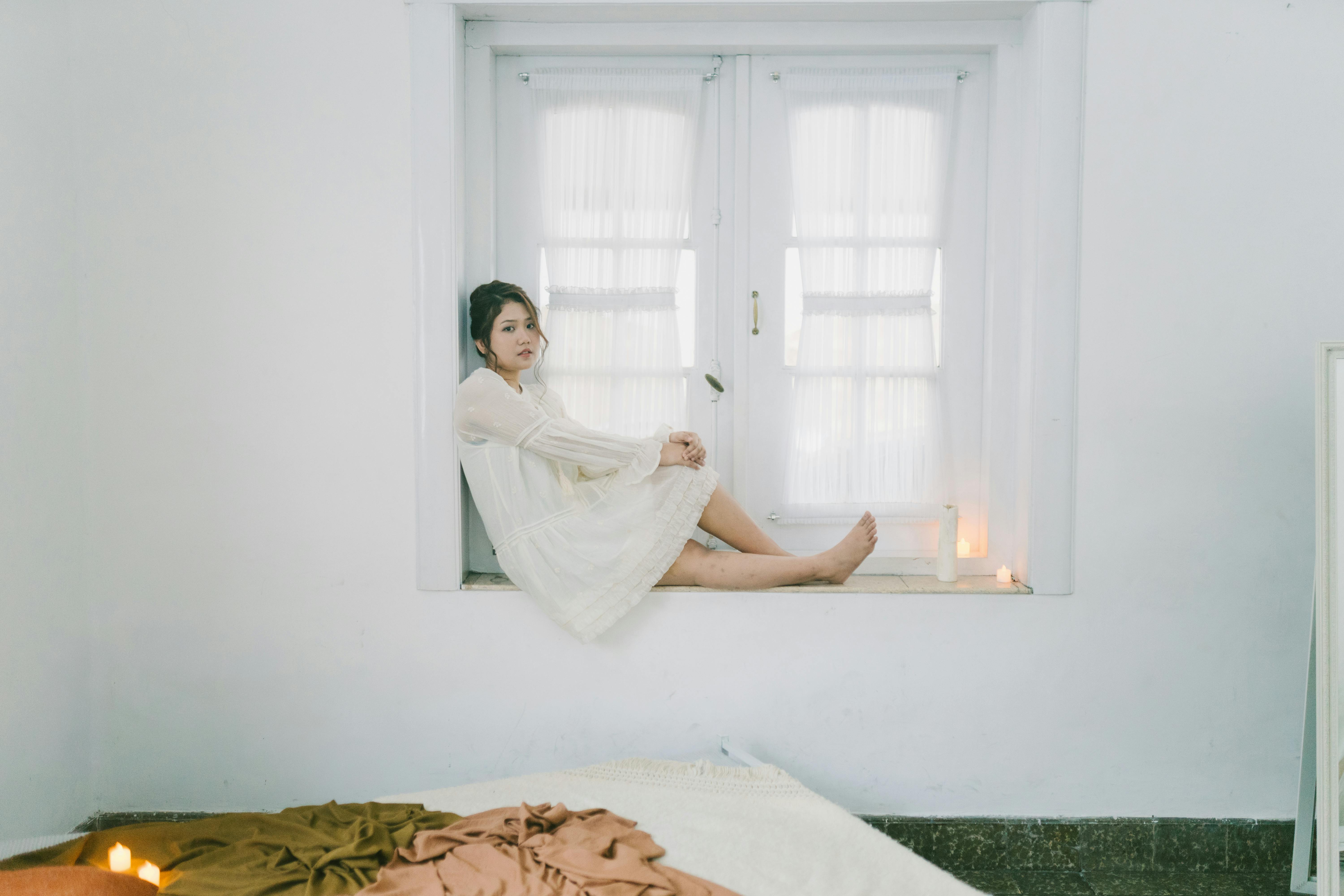 Gratis Fredfyldt portræt af en kvinde, der sidder i en vindueskrog i et hyggeligt soveværelse og omfavner minimalisme. Lagerfoto