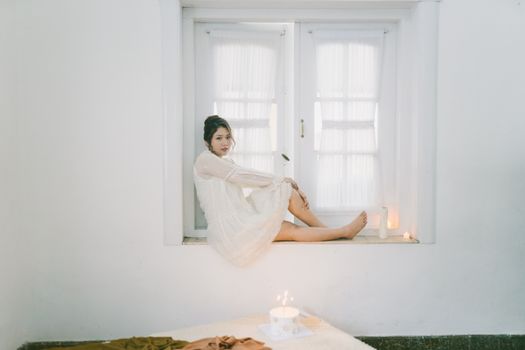 Female sitting on window sill in bright room with candles, creating a soft and serene atmosphere.