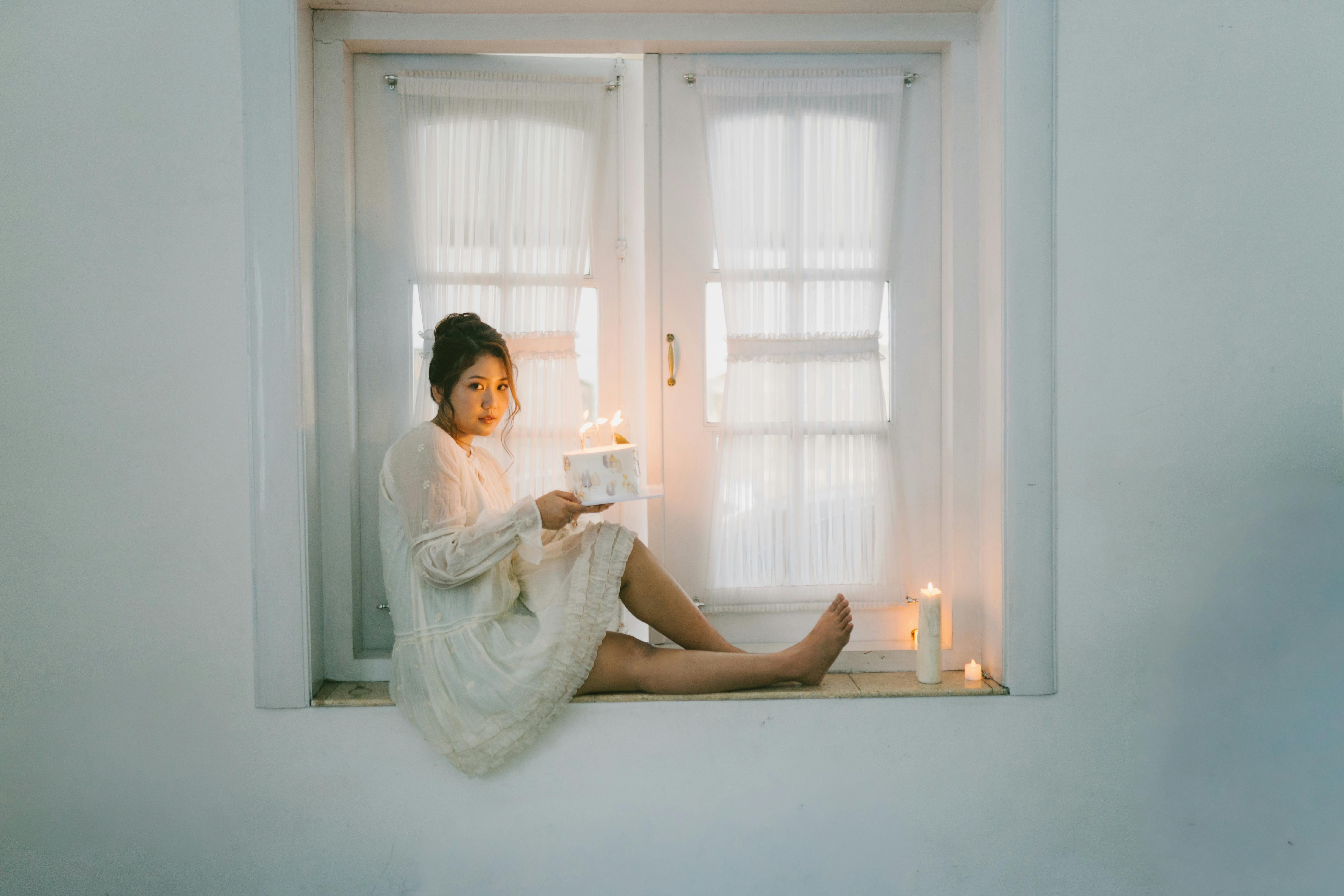 gratis Een vrouw in een witte jurk zit bij een raam met kaarsen, wat een serene en elegante sfeer creëert. Stockfoto