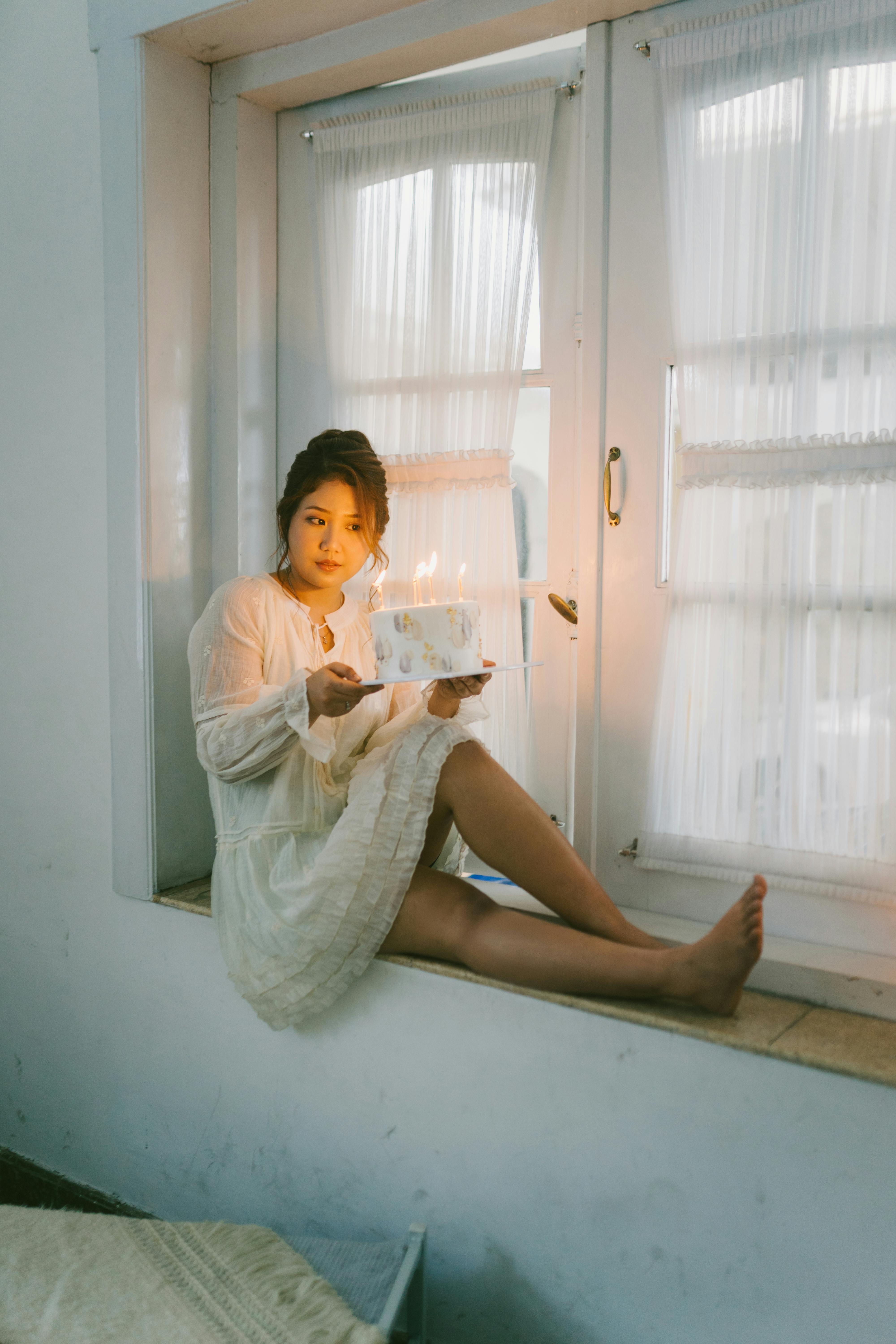 Bezpłatne Spokojna chwila młodej kobiety siedzącej przy oknie i trzymającej tort urodzinowy ze świeczkami. Zdjęcie z galerii