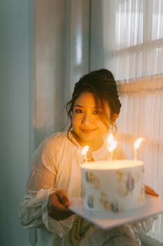 Young woman in a white dress holds a birthday cake with lit candles, creating a warm, celebratory atmosphere.