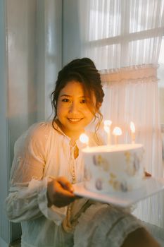 Smiling woman holding birthday cake with candles in cozy indoor setting.