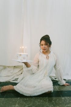 Young woman in white dress holding a birthday cake with candles indoors.