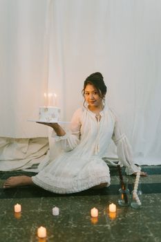 A woman in a white dress holds a birthday cake with candles, surrounded by lit candles indoors.