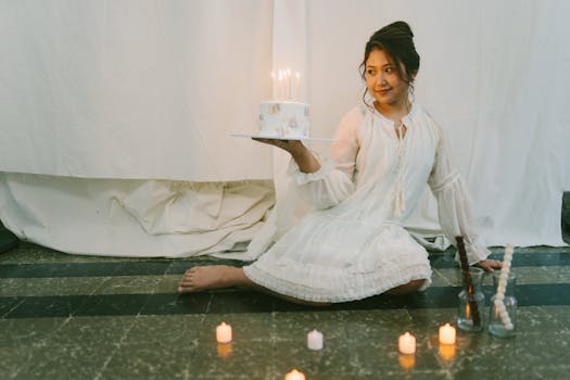 Woman in white dress celebrates birthday with candlelit cake in an intimate setting.