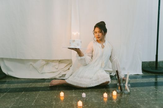 A woman in a white dress holding a birthday cake with candles in an indoor setting.