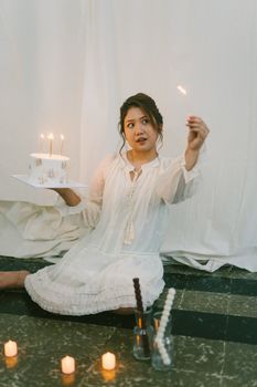 A woman in a white dress holding a cake with lit candles, creating a warm celebratory atmosphere.
