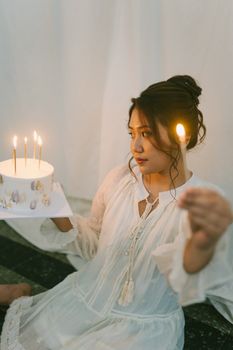 Asian woman in white dress holding birthday cake with lit candles indoors, creating a warm celebratory atmosphere.