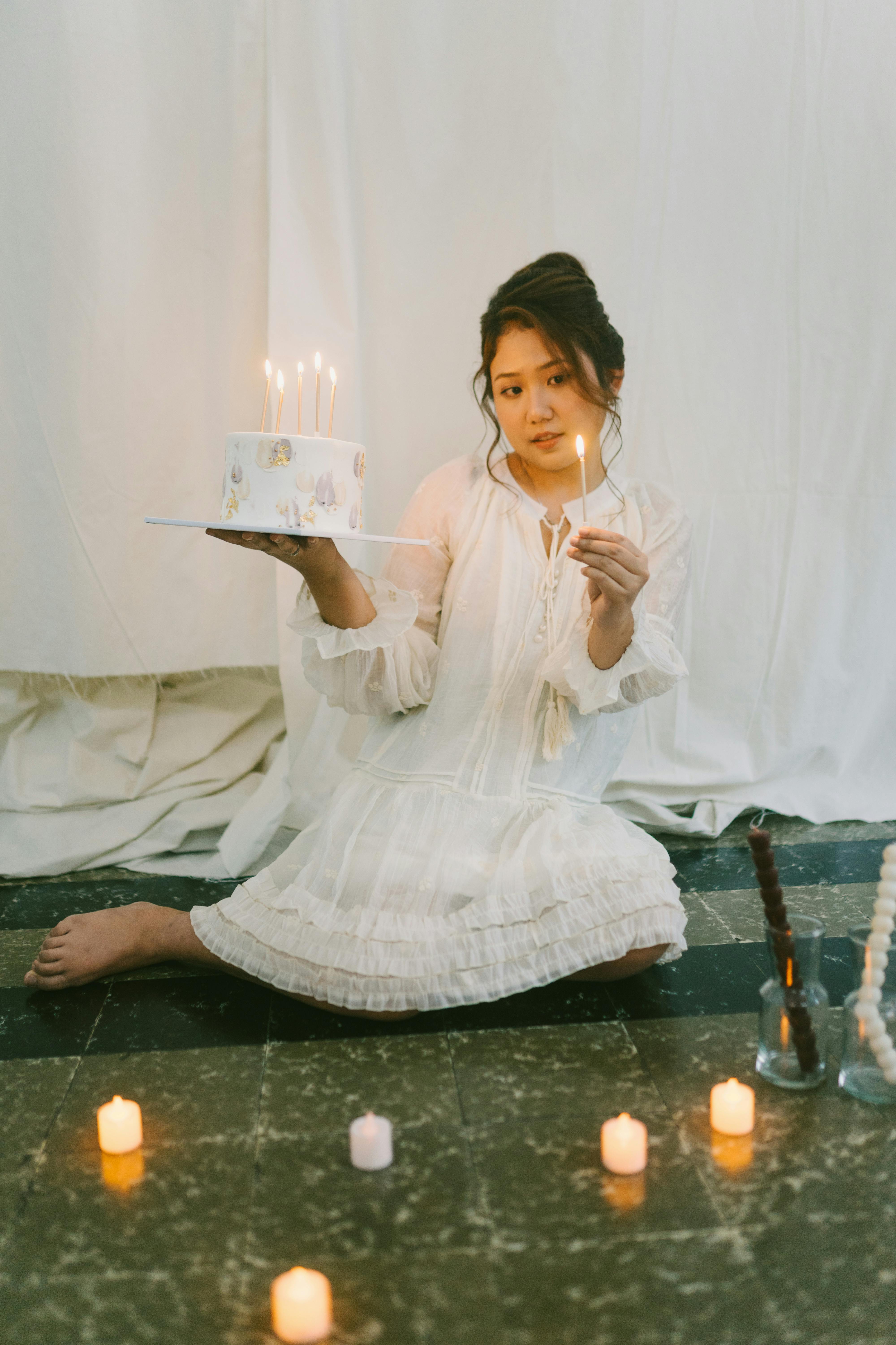 Bezpłatne Młoda kobieta w białej sukni trzyma tort pośród świeczek, co tworzy pogodną urodzinową atmosferę. Zdjęcie z galerii