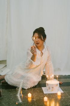 A woman in white dress sits by a cake surrounded by candles, creating a serene ambiance.