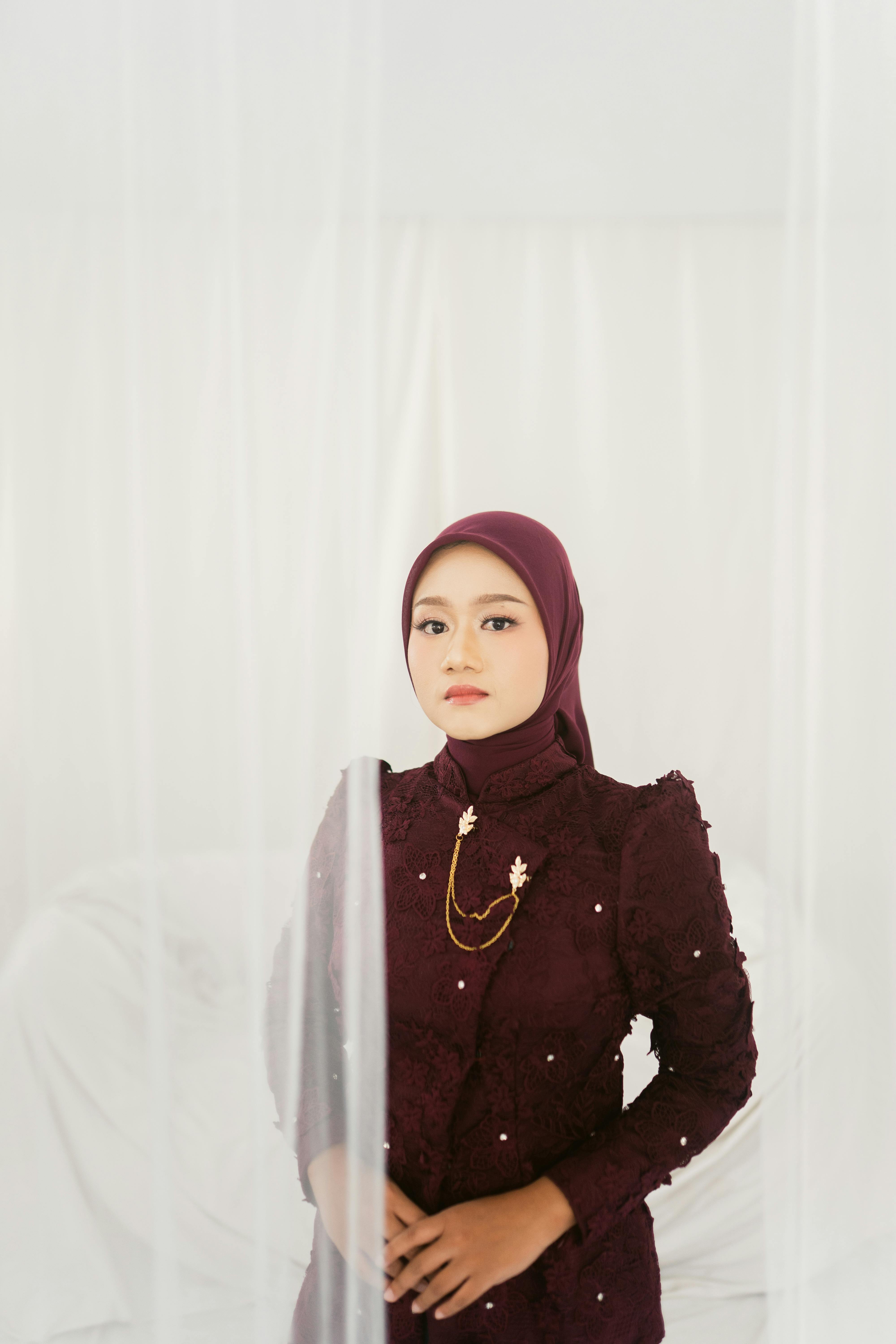 Gratis Potret seorang wanita mengenakan hijab berwarna merah marun dengan ekspresi lembut, berlatar belakang putih yang halus. Foto Stok