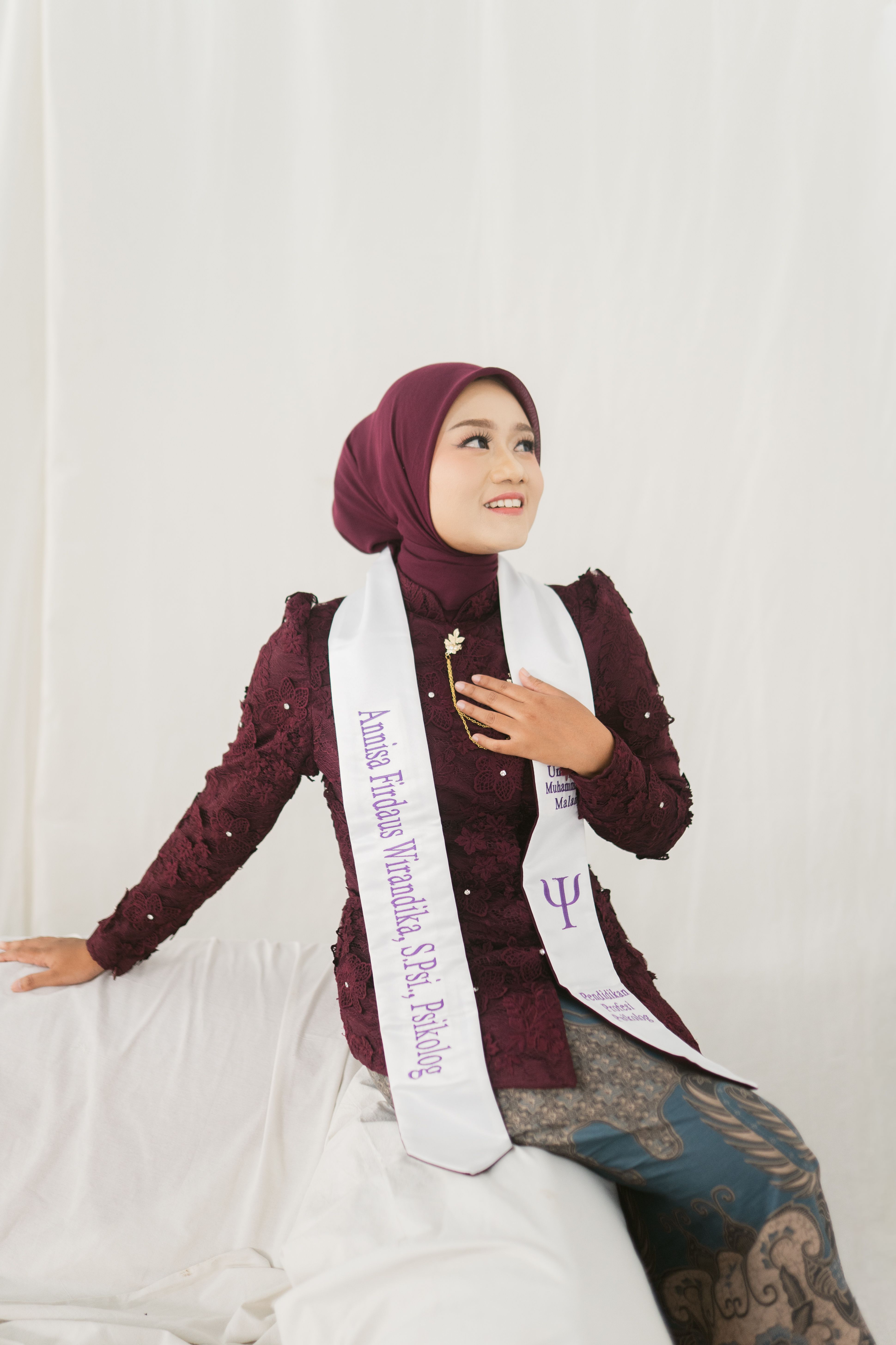 Young woman in maroon attire with a graduation sash, looking joyful indoors.