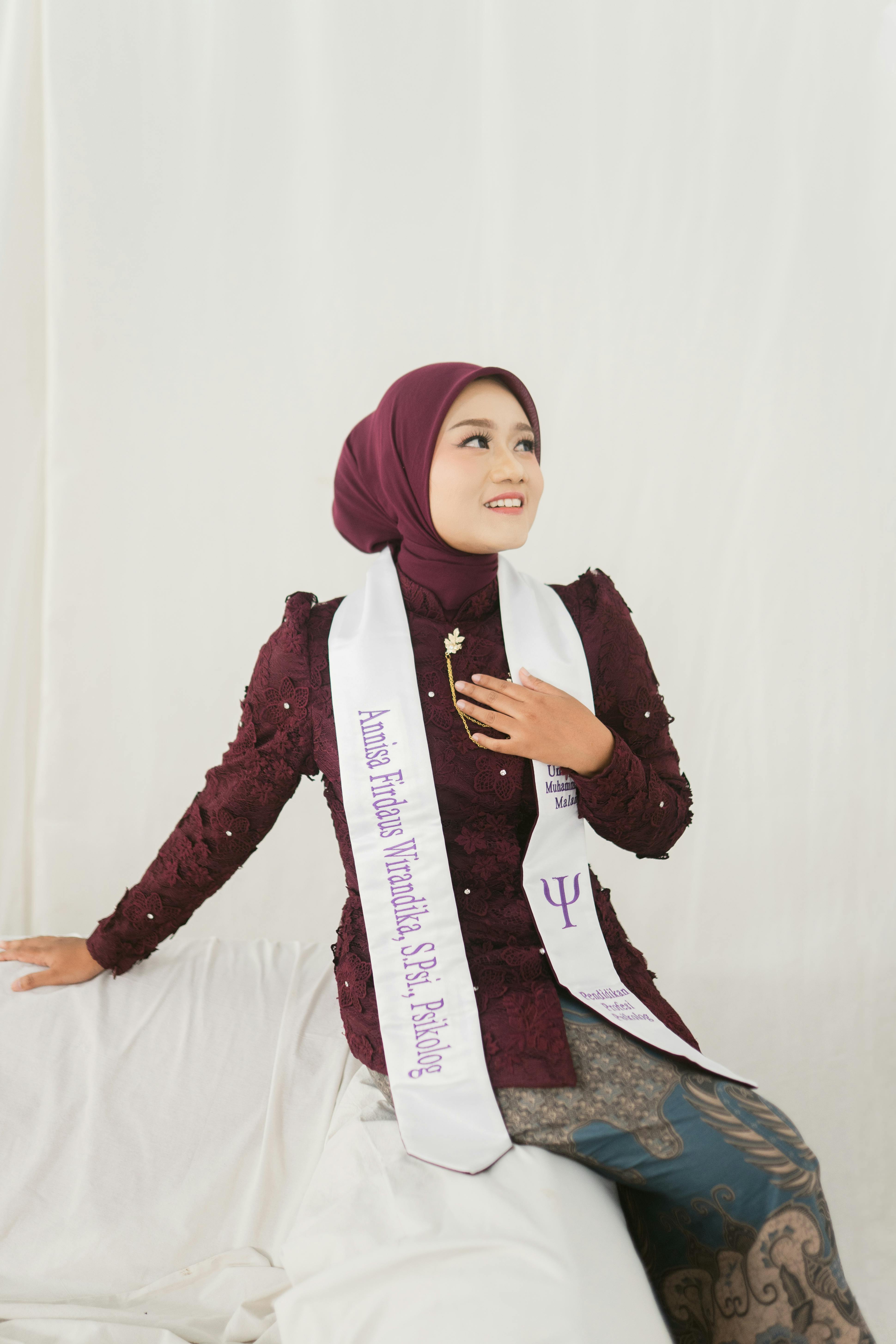 Miễn phí Cô gái trẻ mặc trang phục màu đỏ sẫm, đeo dải băng tốt nghiệp, trông rất vui vẻ khi ở trong nhà. Ảnh lưu trữ