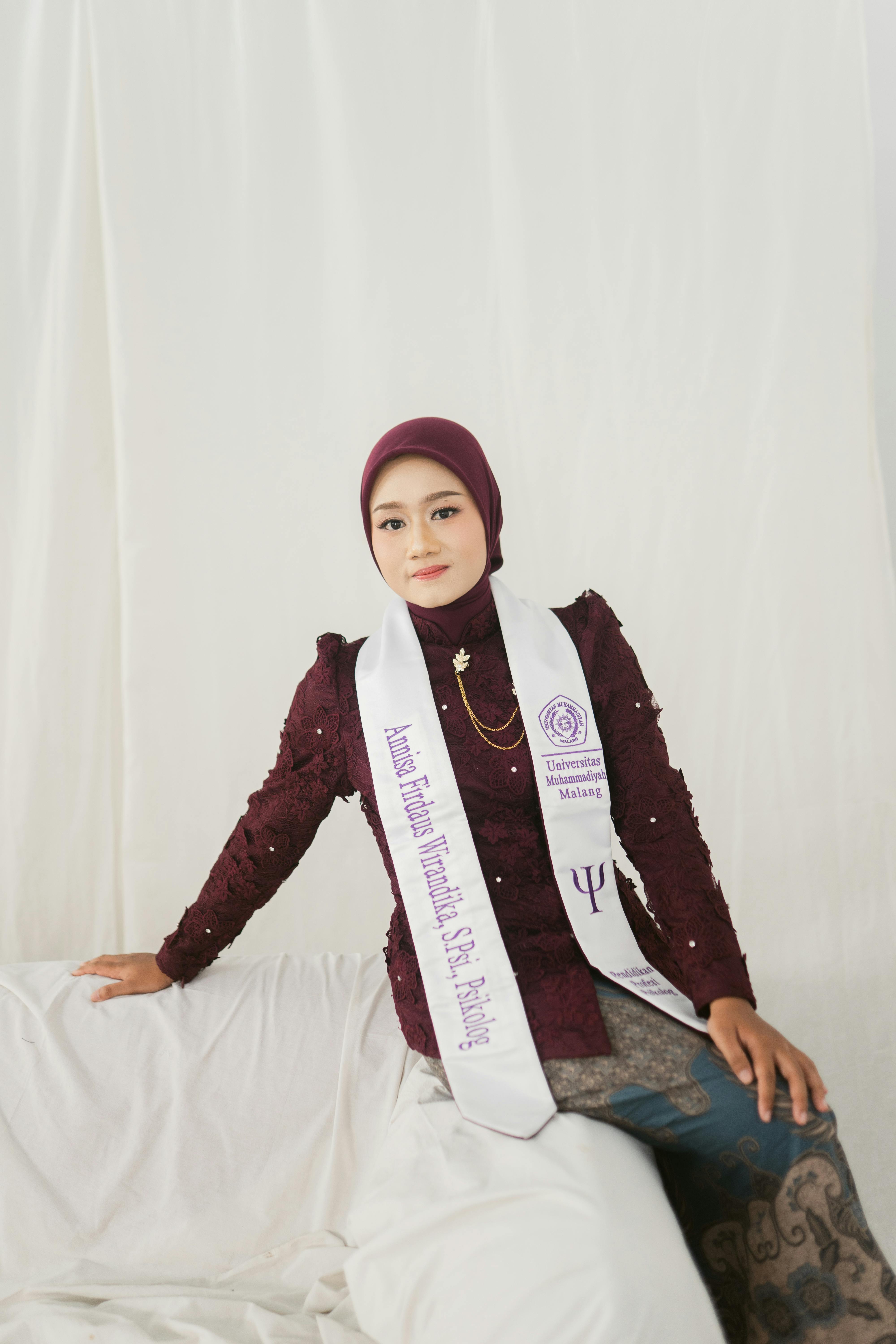 Young woman in maroon traditional attire and graduation sash, seated indoors.