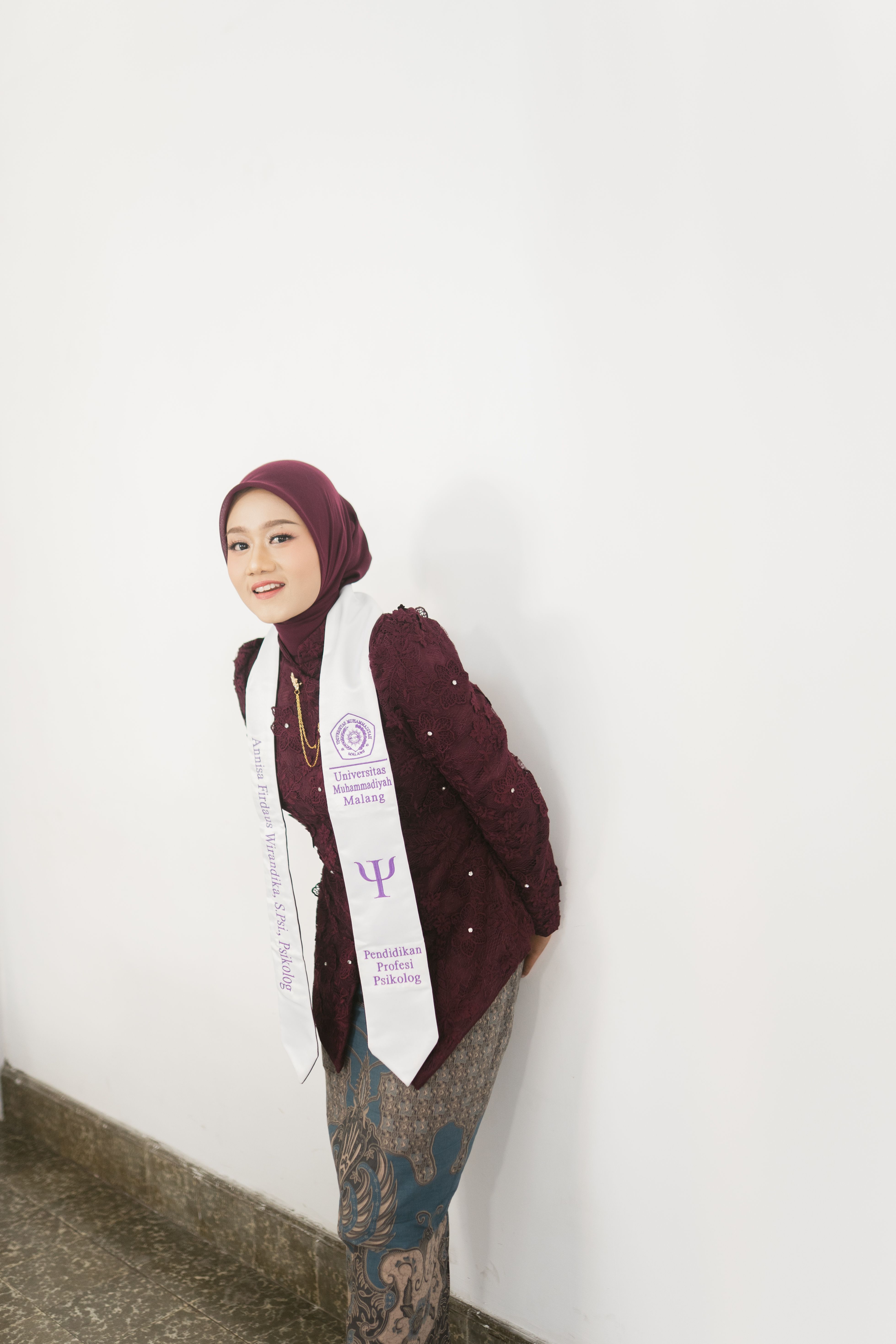A young woman in a maroon hijab and ceremonial sash poses confidently against a white wall indoors.