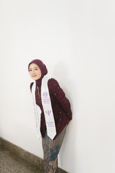 A young woman in a maroon hijab and ceremonial sash poses confidently against a white wall indoors. - Fotografer Sidoarjo