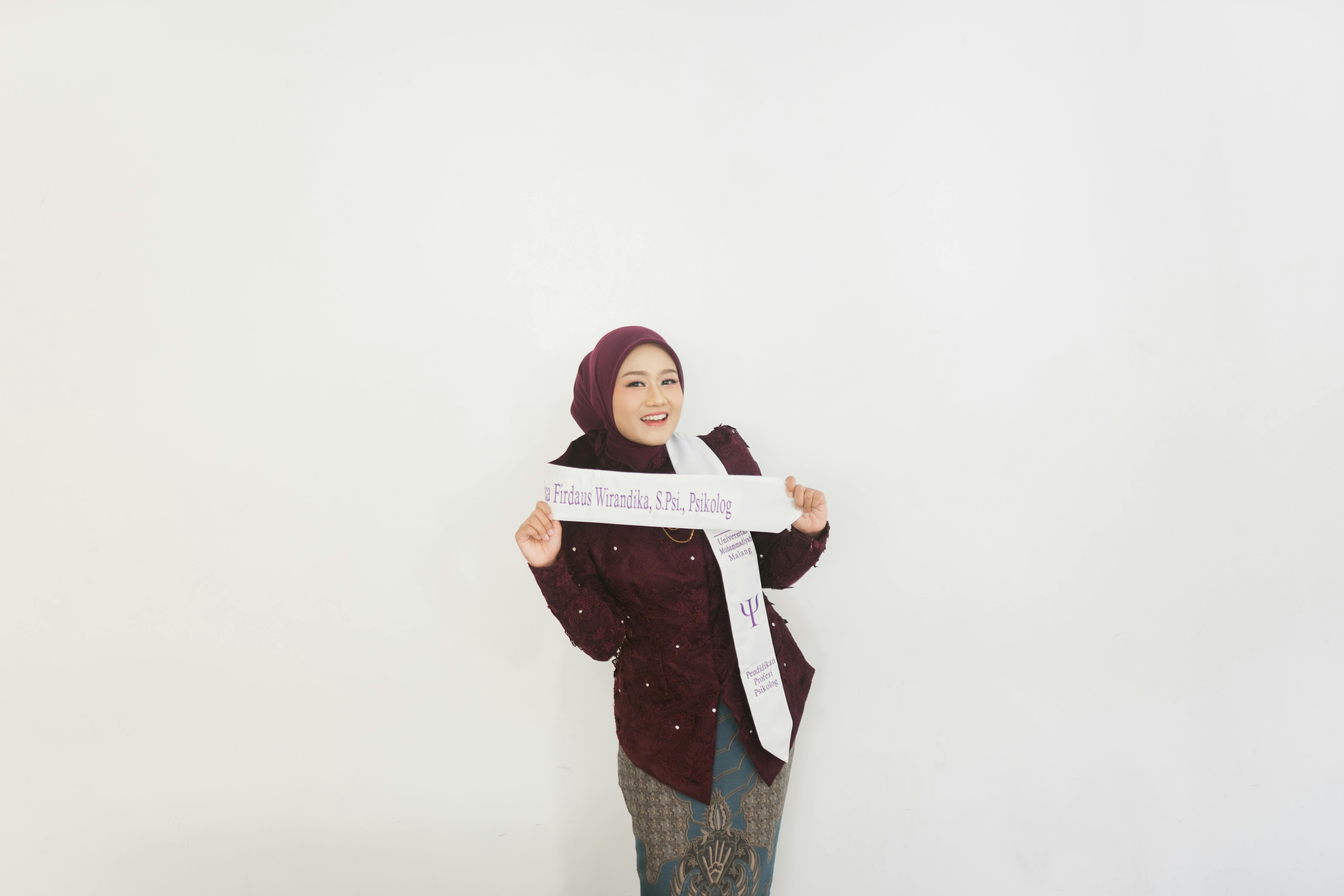 Young woman in a maroon outfit celebrating her graduation with a sash, smiling against a white background.