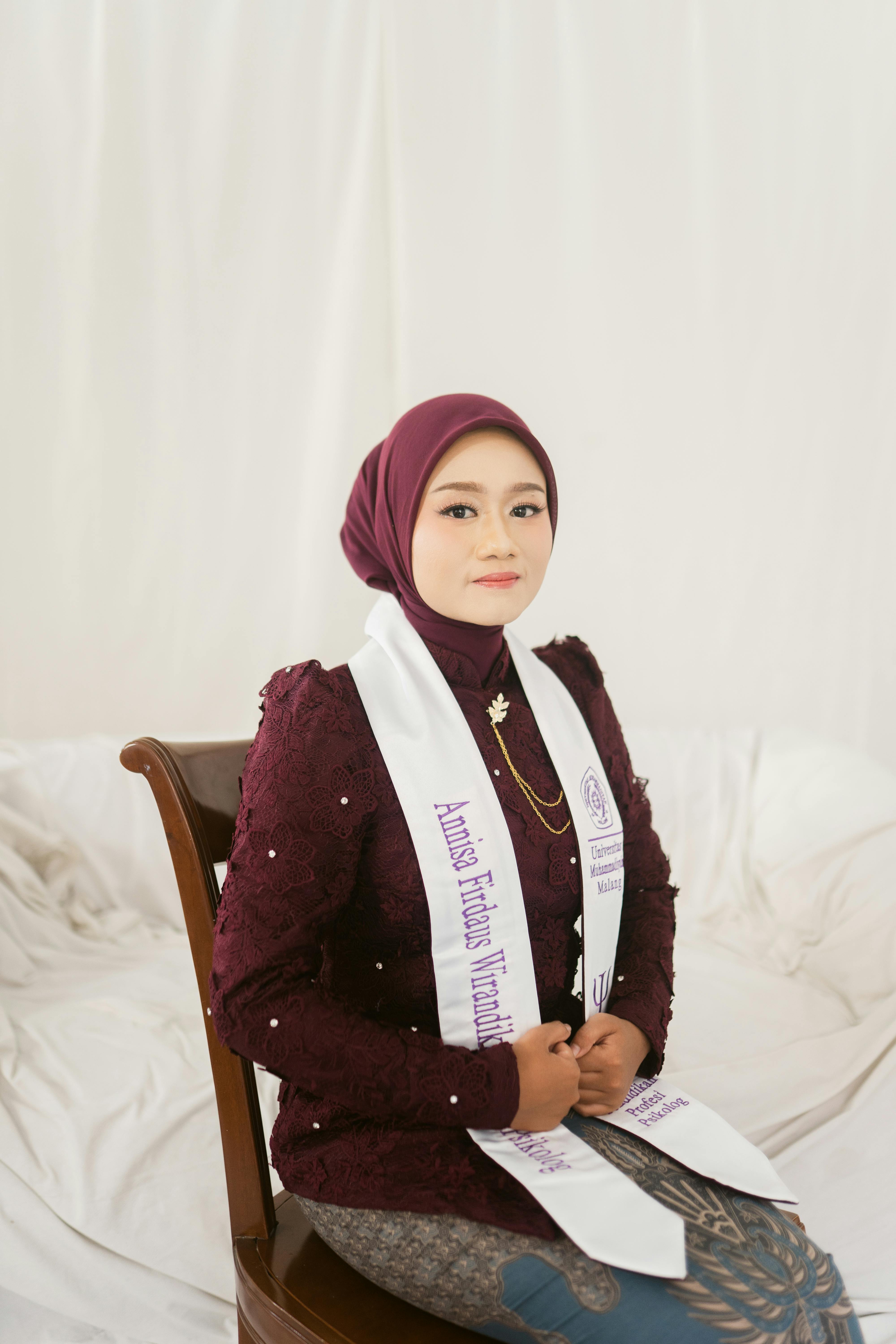 Elegant portrait of a young woman wearing a hijab and graduation stole, seated indoors.