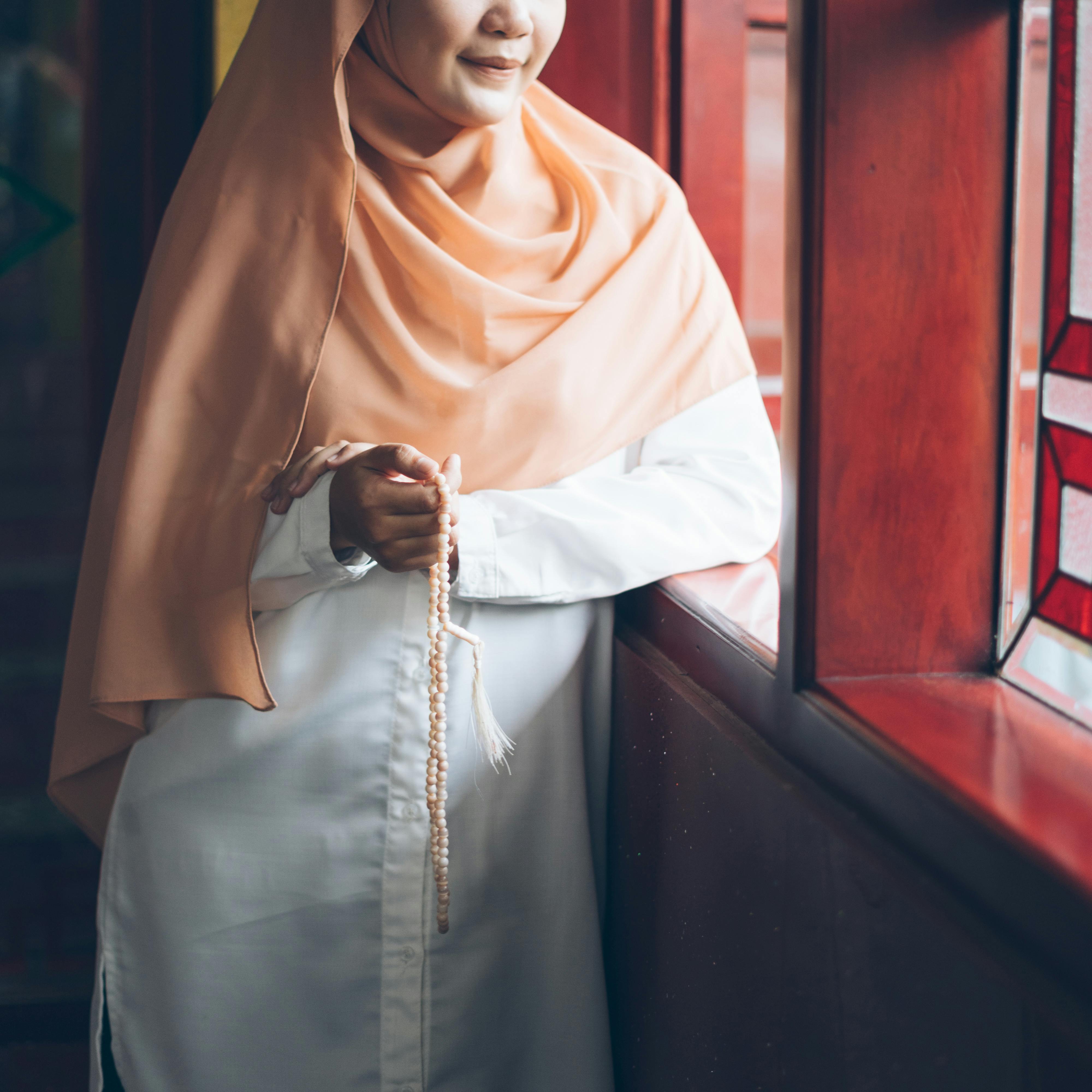 Miễn phí Một người phụ nữ Hồi giáo đội khăn trùm đầu đứng bên cửa sổ, tay cầm tràng hạt, trầm tư suy nghĩ trong không gian nội thất yên bình. Ảnh lưu trữ
