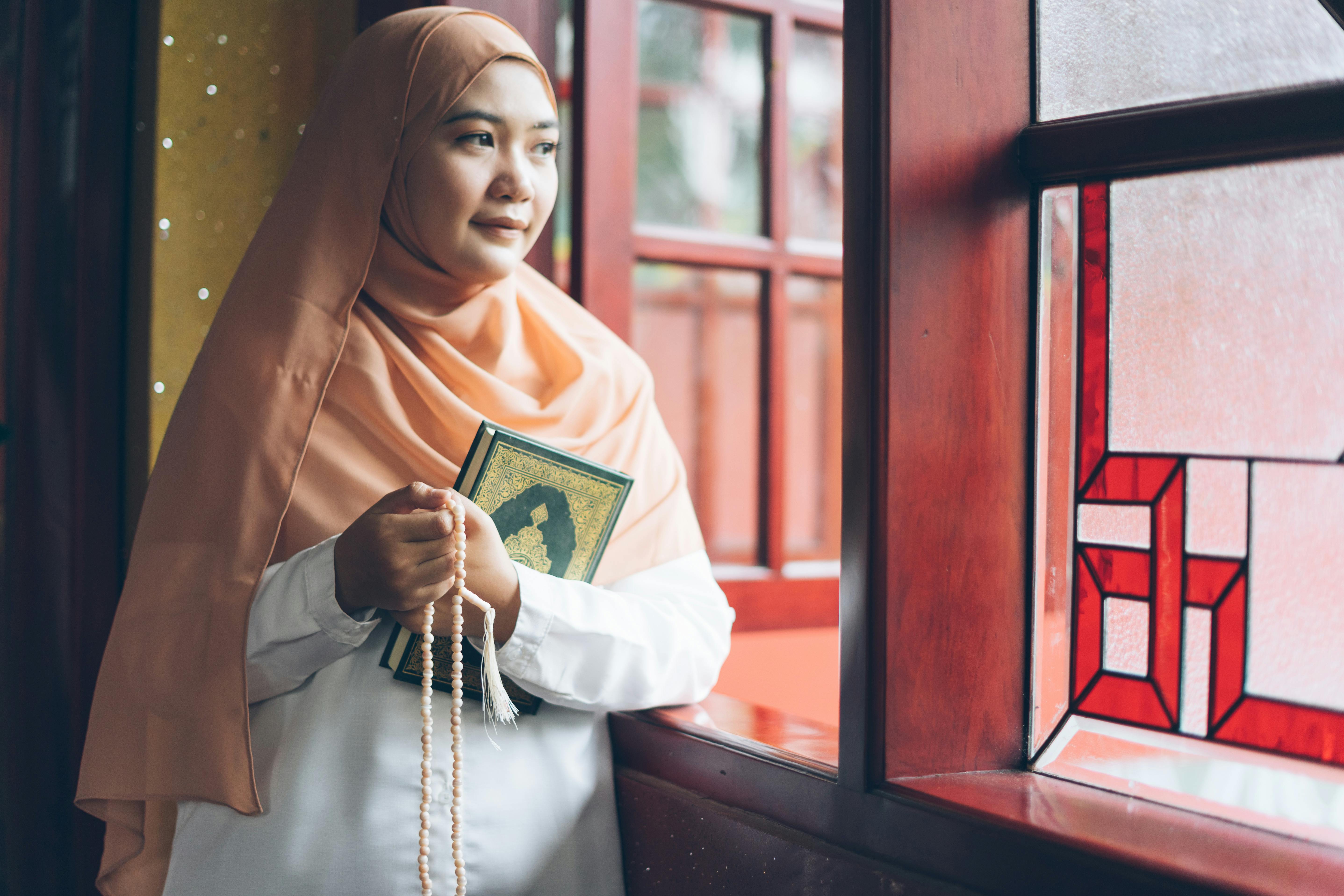 Miễn phí Một người phụ nữ đội khăn trùm đầu cầm kinh Quran và tràng hạt bên cửa sổ kính màu, toát lên vẻ bình yên và trầm tư. Ảnh lưu trữ