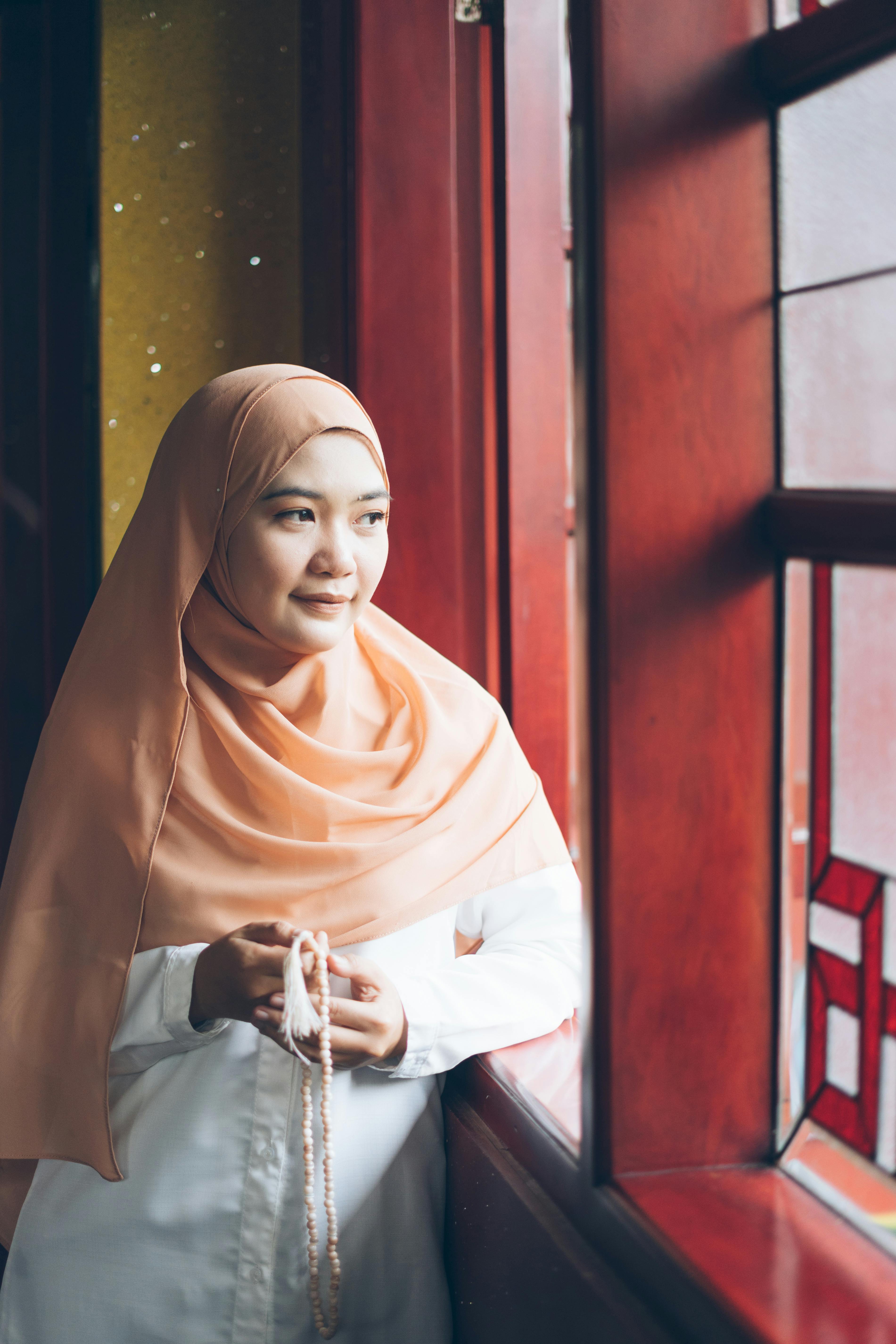 Bezpłatne Kobieta w brzoskwiniowym hidżabie trzyma różaniec i patrzy przez witraż. Zdjęcie z galerii