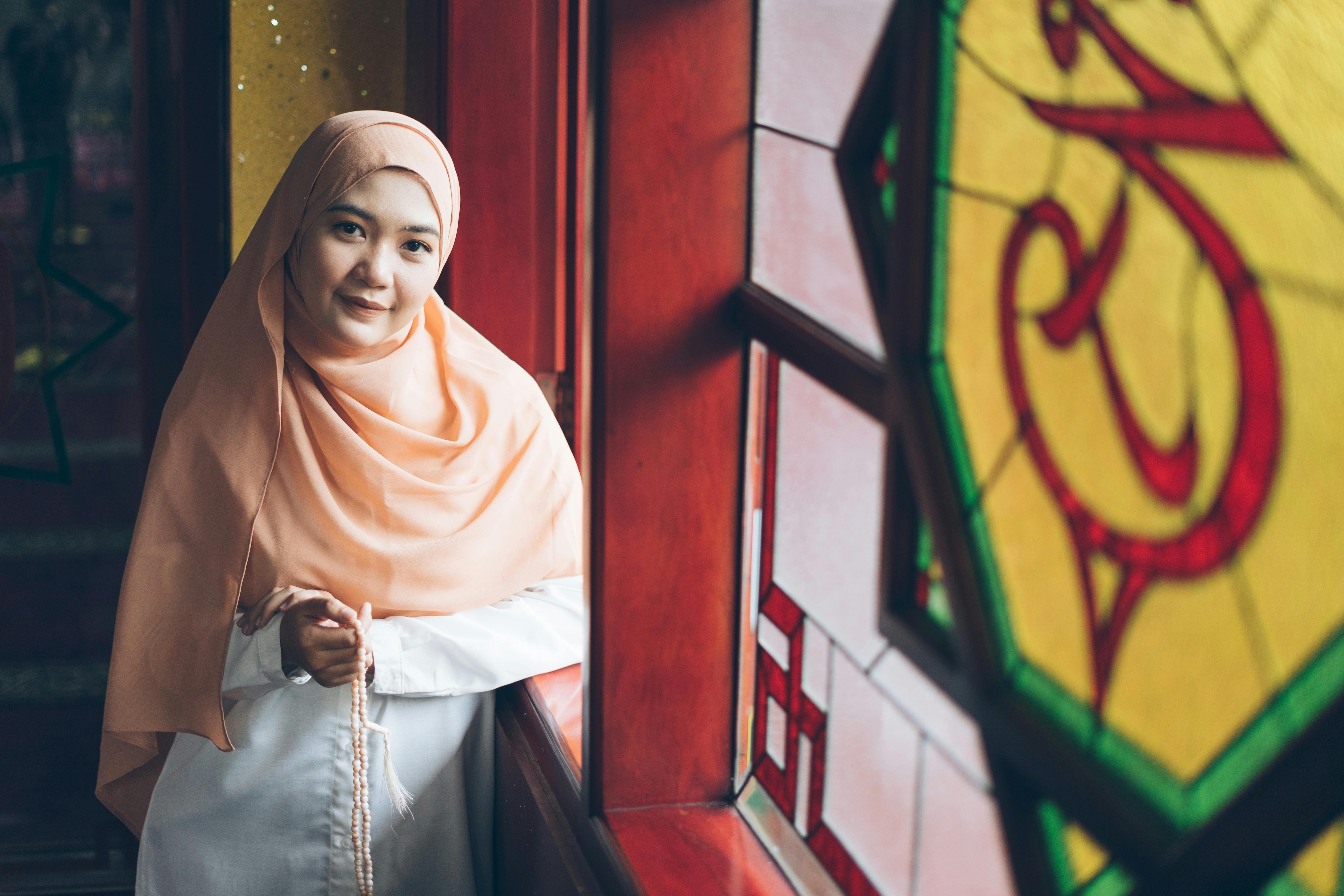 Bezpłatne Kobieta w brzoskwiniowym hidżabie, trzymająca koraliki modlitewne w pobliżu kolorowych witraży w pomieszczeniu zamkniętym. Zdjęcie z galerii