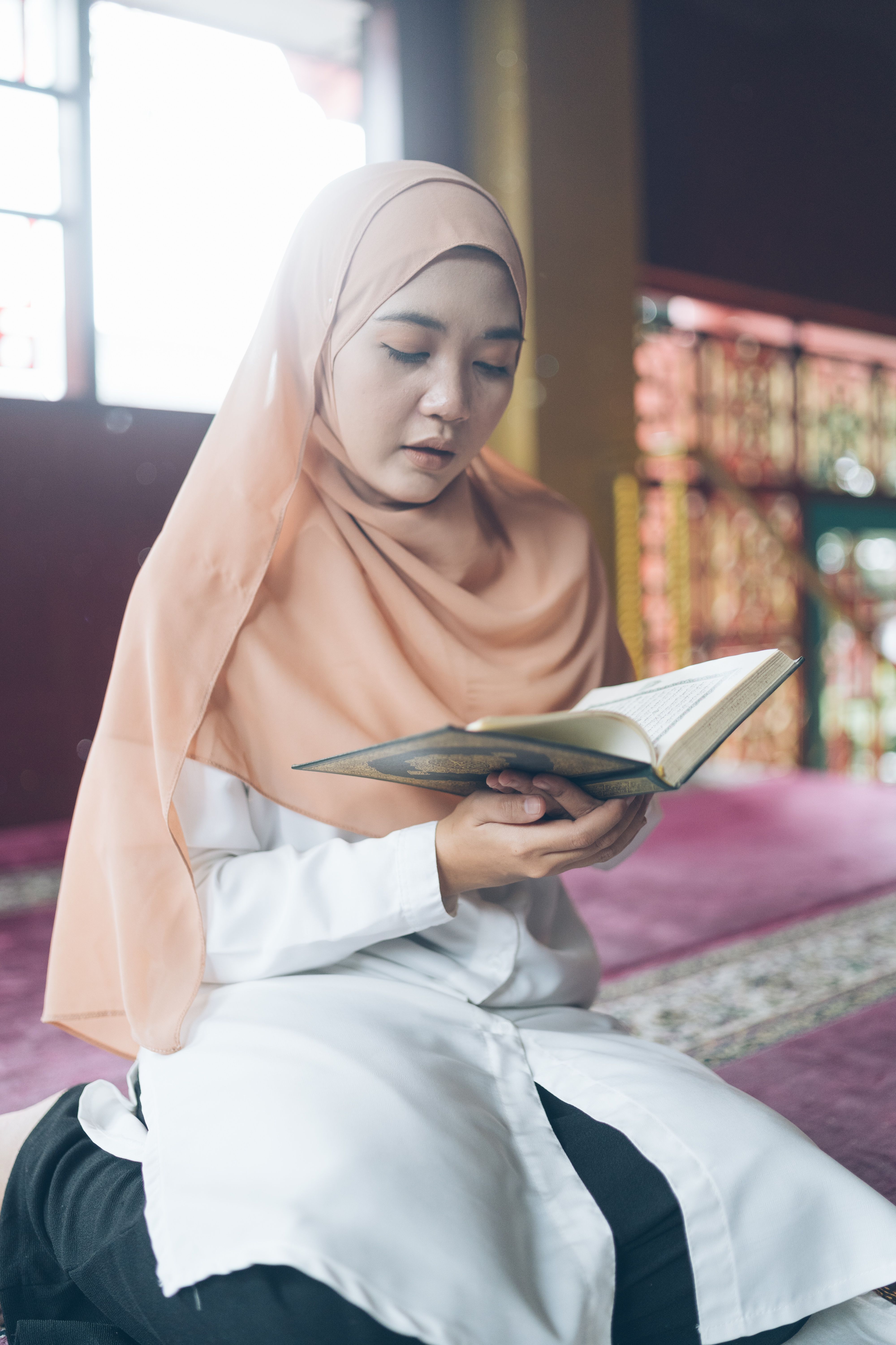 A young woman wearing a hijab reads a book in a mosque, reflecting devotion.