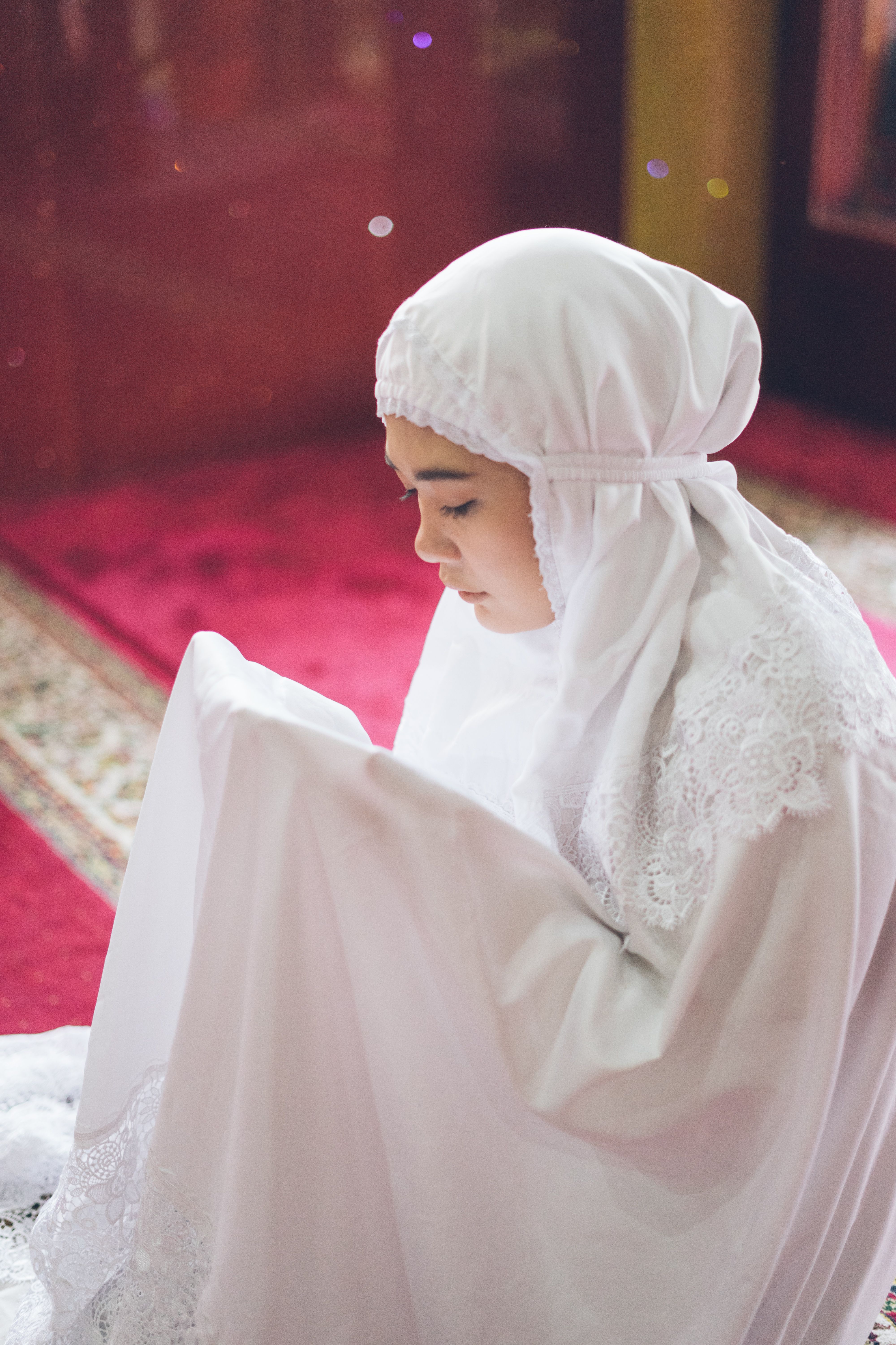 A young woman donning a white traditional hijab, engaged in quiet prayer indoors.