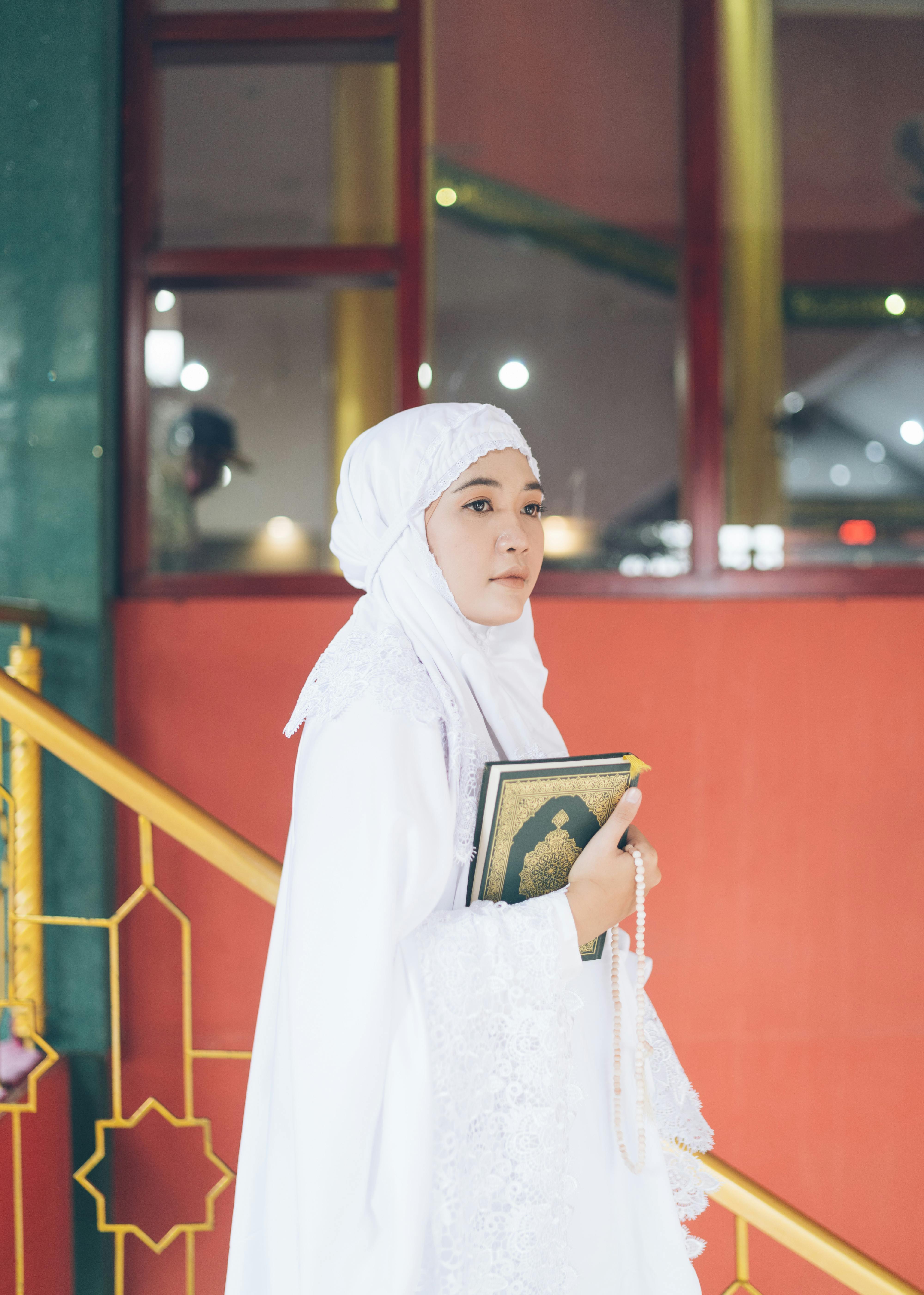 Gratis En ung kvinde i en hvid hijab holder Koranen og bedepæle inde i en moské, hvilket afspejler spiritualitet. Lagerfoto